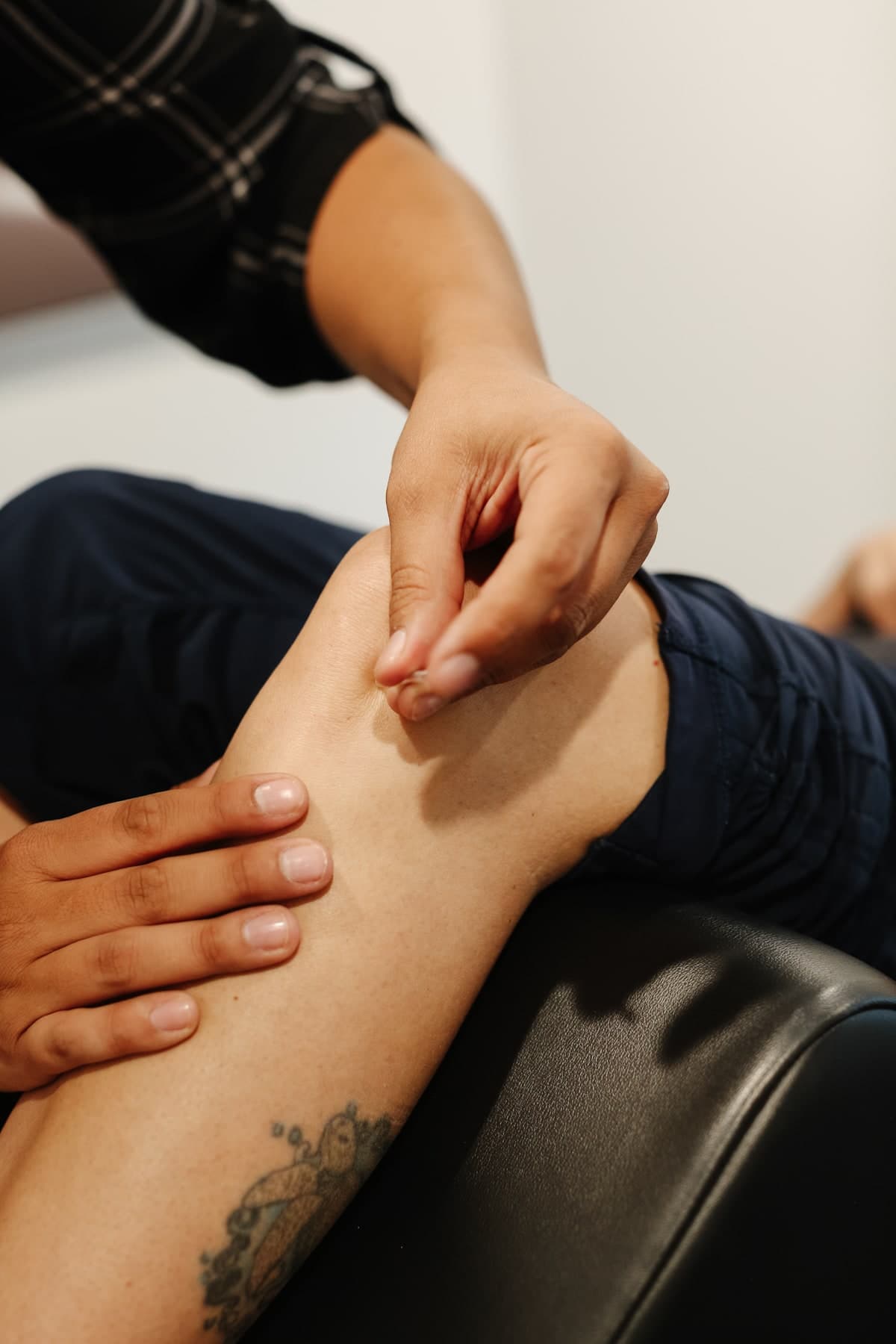 Close-up of a physiotherapist's hands performing a manual treatment on a patient's knee and lower leg while the patient lies on a black leather treatment table, patient wearing navy pants and a tattoo visible on the lower leg, Swift Current