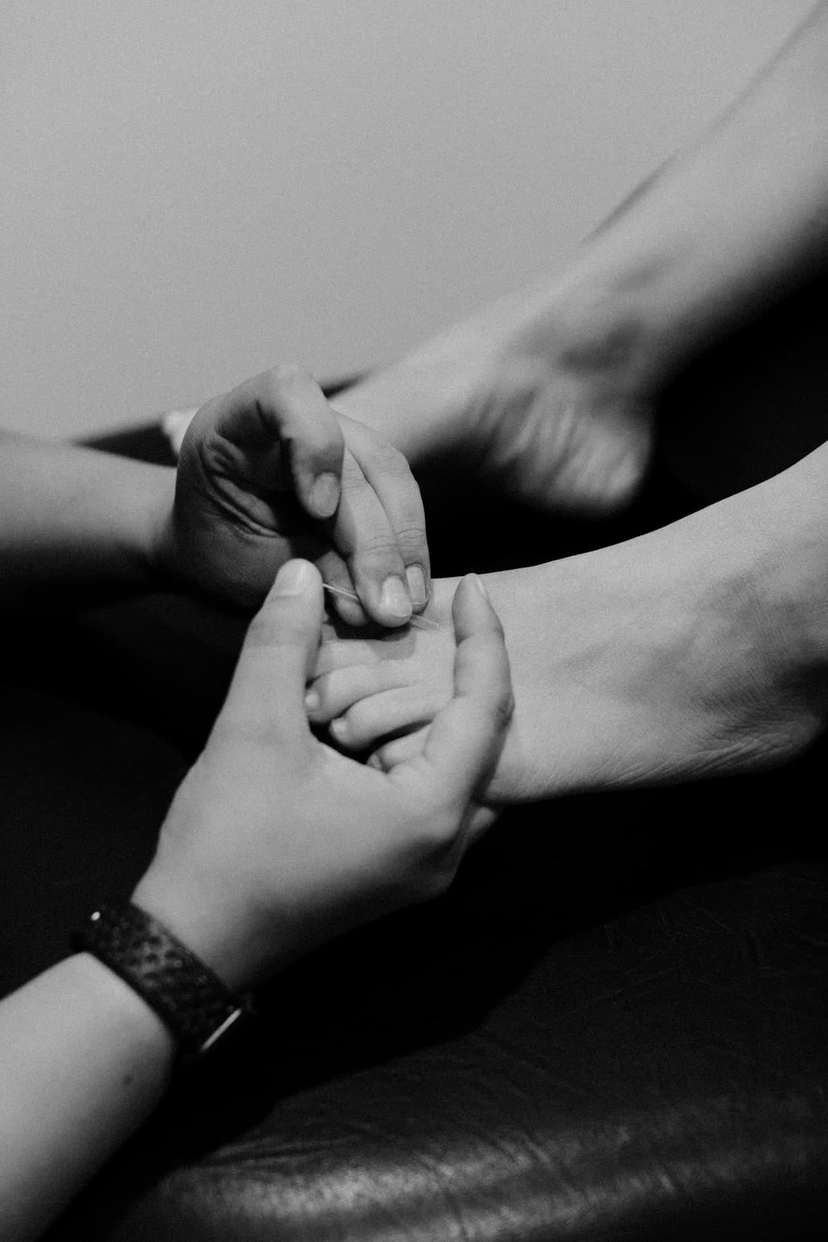 Black and white close-up of a physiotherapist's hands holding a patient's hand, with a fine acupuncture needle being inserted between the fingers, patient's forearm and foot visible in the background, Swift Current