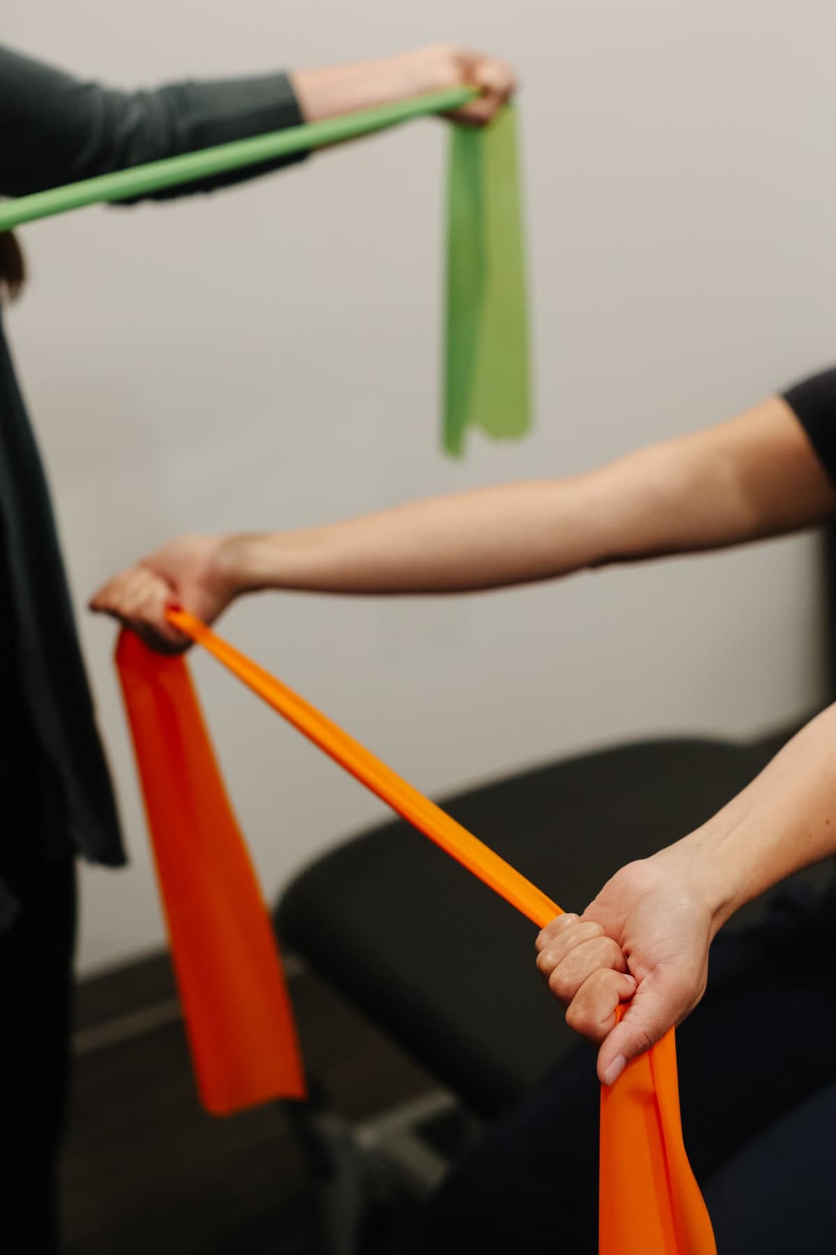 Two hands stretching green and orange resistance bands in a clinic setting, a person in dark clothing on the left and a person in dark clothing on the right, a black treatment bench in the background, Swift Current