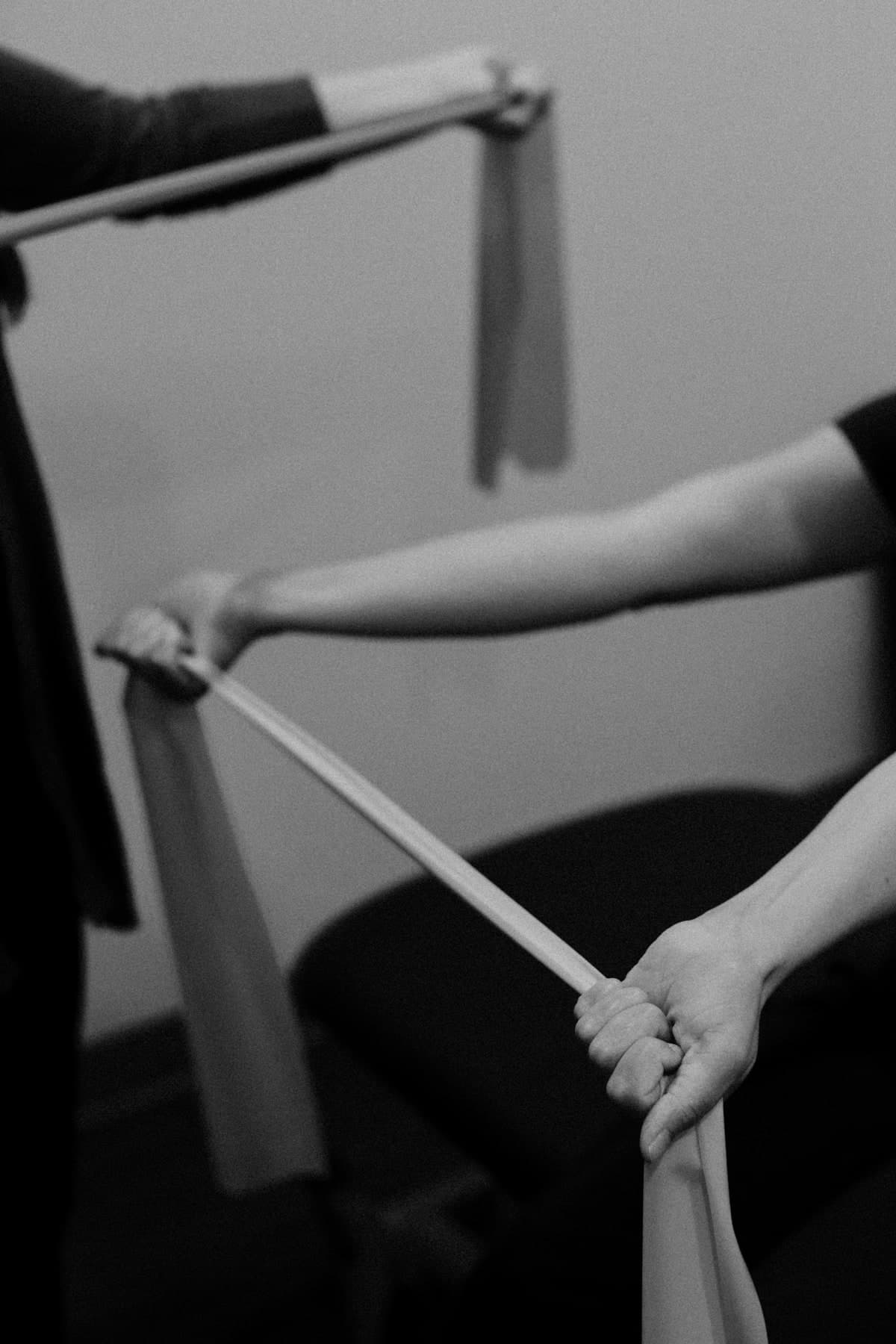 Black and white photo of two people's hands and forearms stretching a resistance band between them in a clinic setting, Swift Current