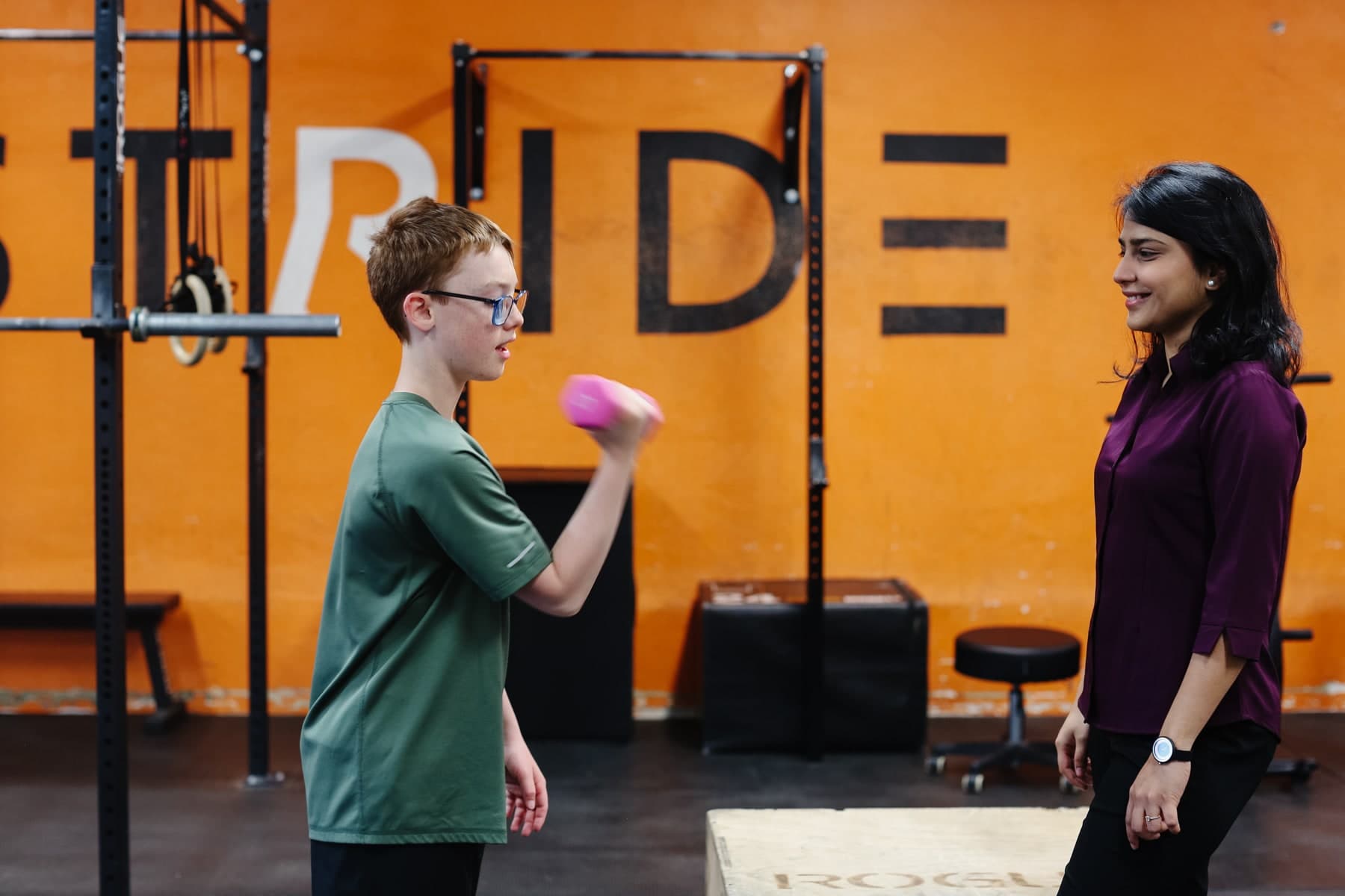 A young boy in glasses and a green t-shirt lifting a pink dumbbell while a smiling woman in a purple blouse watches him in a gym with an orange wall