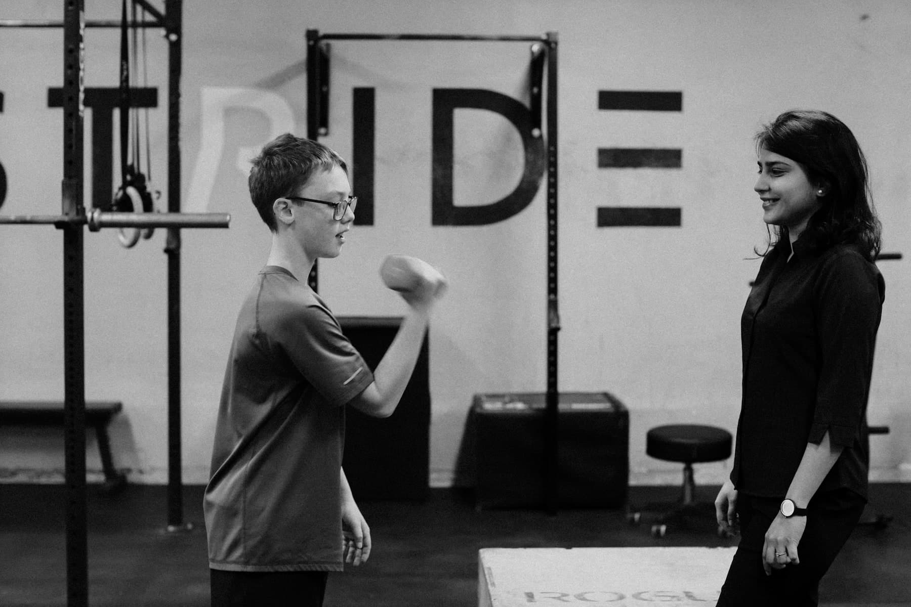 Black and white photo of a young boy in glasses doing a dumbbell curl while a smiling woman in a dark shirt watches, in a gym with 'PRIDE' lettering on the wall, Swift Current
