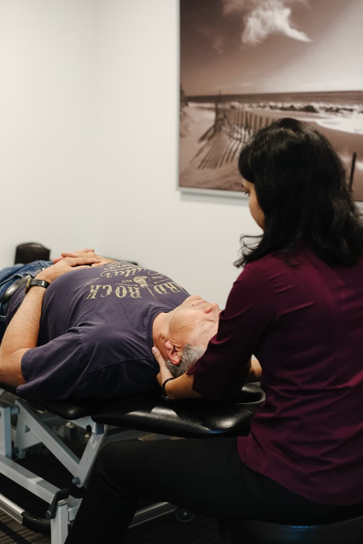 Physiotherapist in a maroon top supporting the head and neck of a patient lying face up on a treatment table, with a black-and-white beach photograph on the wall behind them, Swift Current