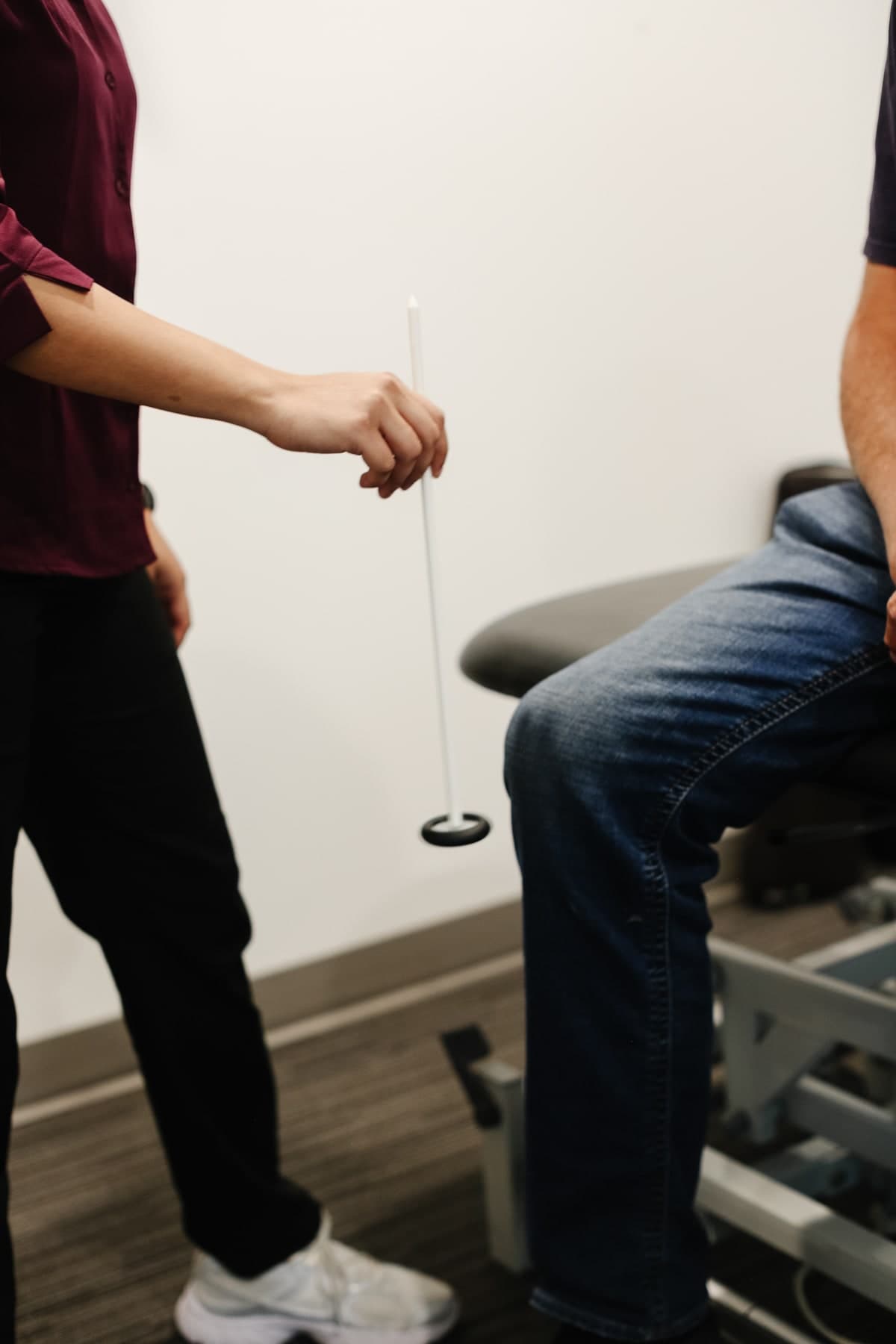 Physiotherapist in a maroon shirt and black pants holding a white rod with a black weighted base, a seated patient in jeans beside a treatment table, Swift Current