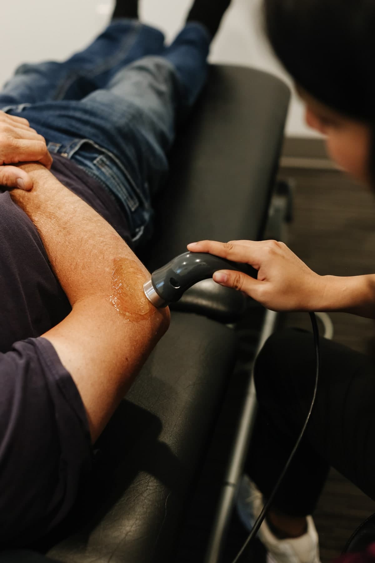 Close-up of a physiotherapist in black clothing holding a grey handheld ultrasound or shockwave device with a silver tip against the outer thigh of a patient lying on a black treatment table, with a clear gel visible on the skin