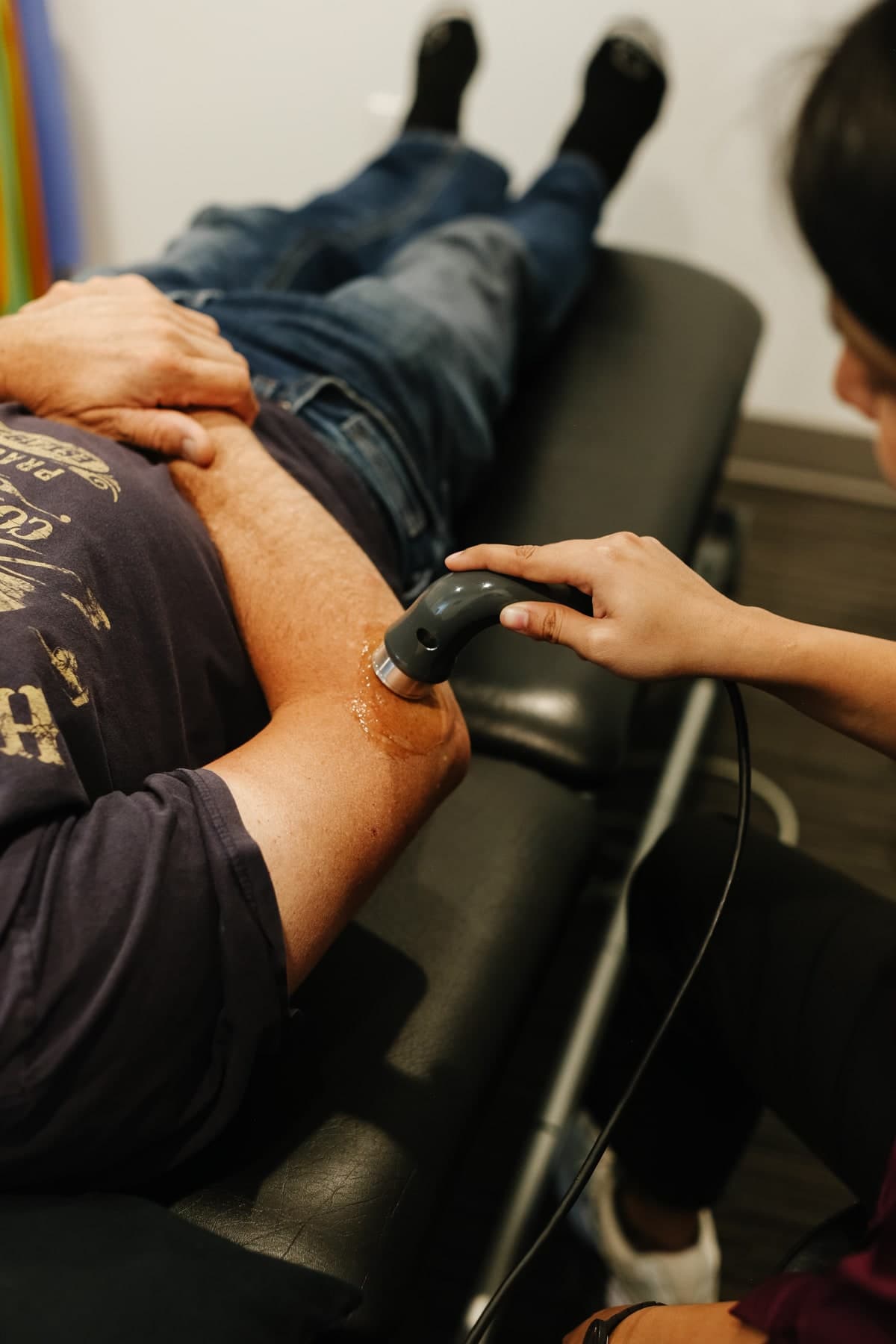 Patient lying on a treatment table in jeans and dark socks while a practitioner in a maroon top applies a handheld shockwave therapy device with gel to the outer thigh