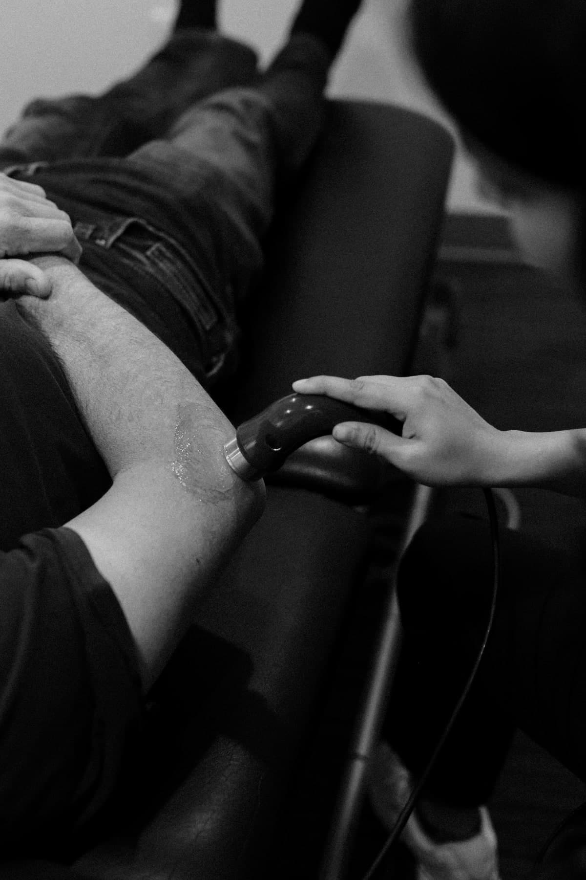 Black and white photo of a physiotherapist holding a shockwave therapy applicator against the outer thigh of a seated patient in jeans, with the patient's hands resting on the knee