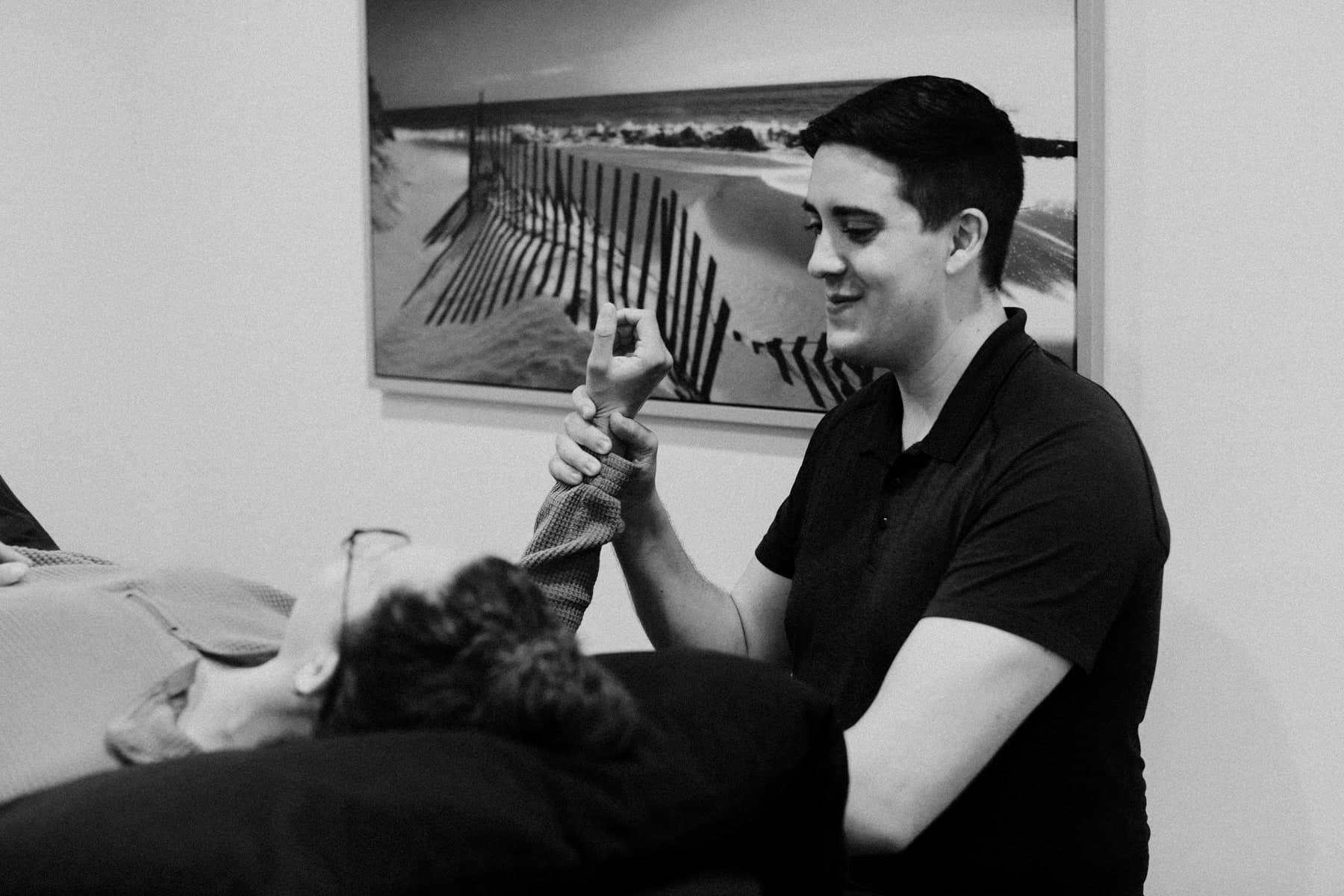 Black and white photo of a physiotherapist in a dark polo shirt smiling while gently guiding a patient's wrist and hand, the patient lying on a treatment table with face partly visible, a framed beach photograph with wooden snow fencing on the wall behind, Swift Current