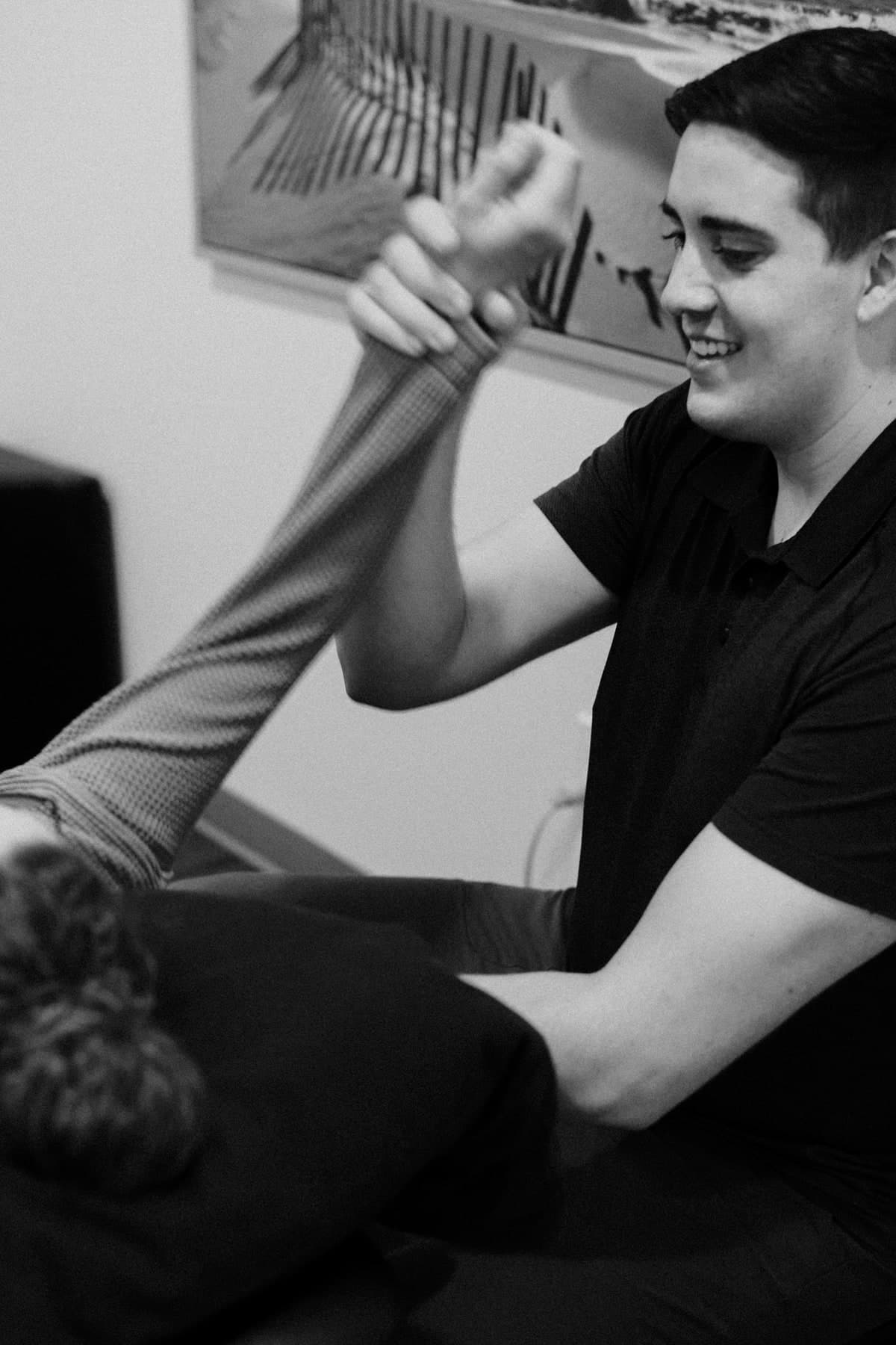 Black and white photo of a smiling physiotherapist in a dark polo shirt holding a patient's leg raised and bent at the knee while the patient lies on a treatment table, with a framed artwork on the wall behind