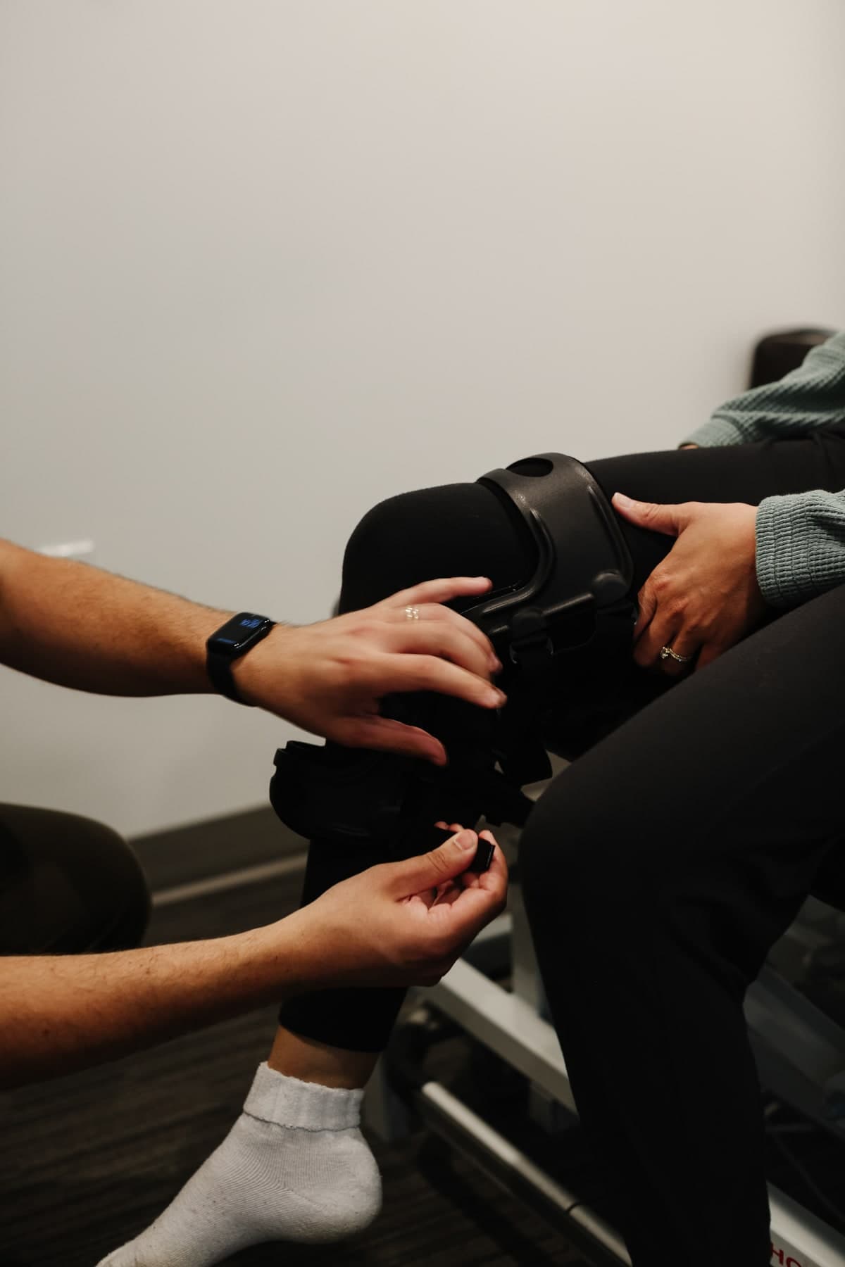 Physiotherapist in a grey shirt fitting a black hinged knee brace on a seated patient in black leggings and white socks, in a clinic room with a grey wall and dark floor in Swift Current