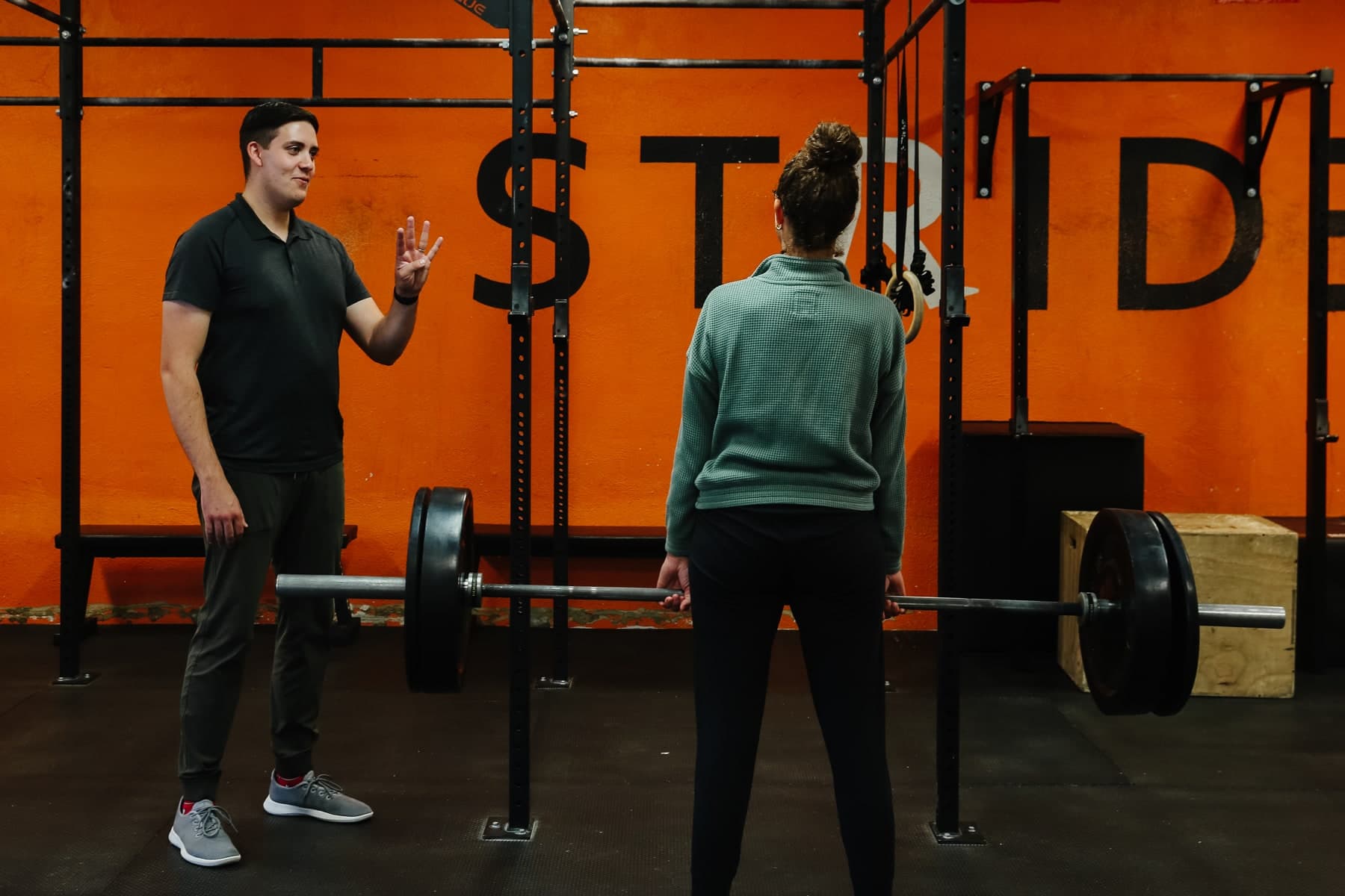 Physiotherapist in a grey polo and grey sneakers gesturing with three fingers while a woman with her hair in a bun, wearing a green sweater and black leggings, holds a loaded barbell in a gym with orange walls and black squat racks in Swift Current