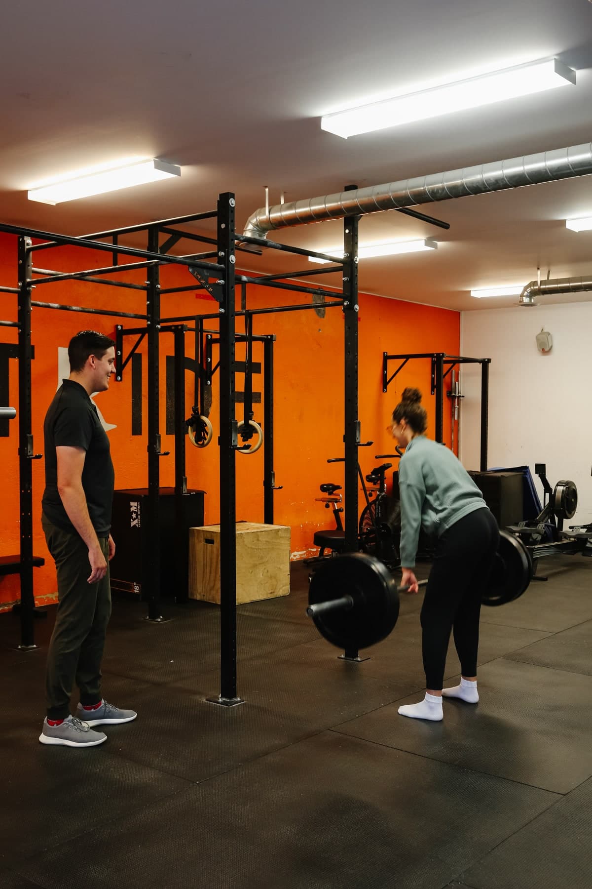 Man in a black polo and grey joggers watching a woman in a grey sweater and black leggings lift a loaded barbell in a training gym with an orange wall, pull-up rigs and rings, and a wooden box in the background, Swift Current
