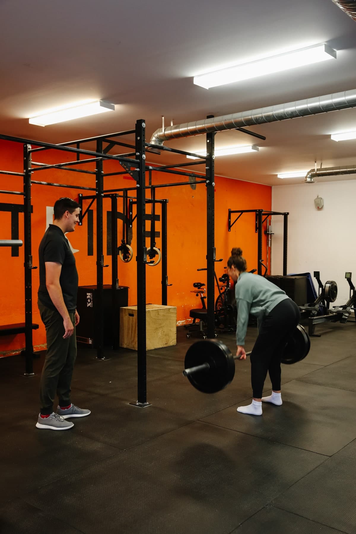 Two people in a gym with orange walls and black squat racks; a man in a black polo stands watching while a woman in a grey top and black leggings holds a loaded barbell at hip height