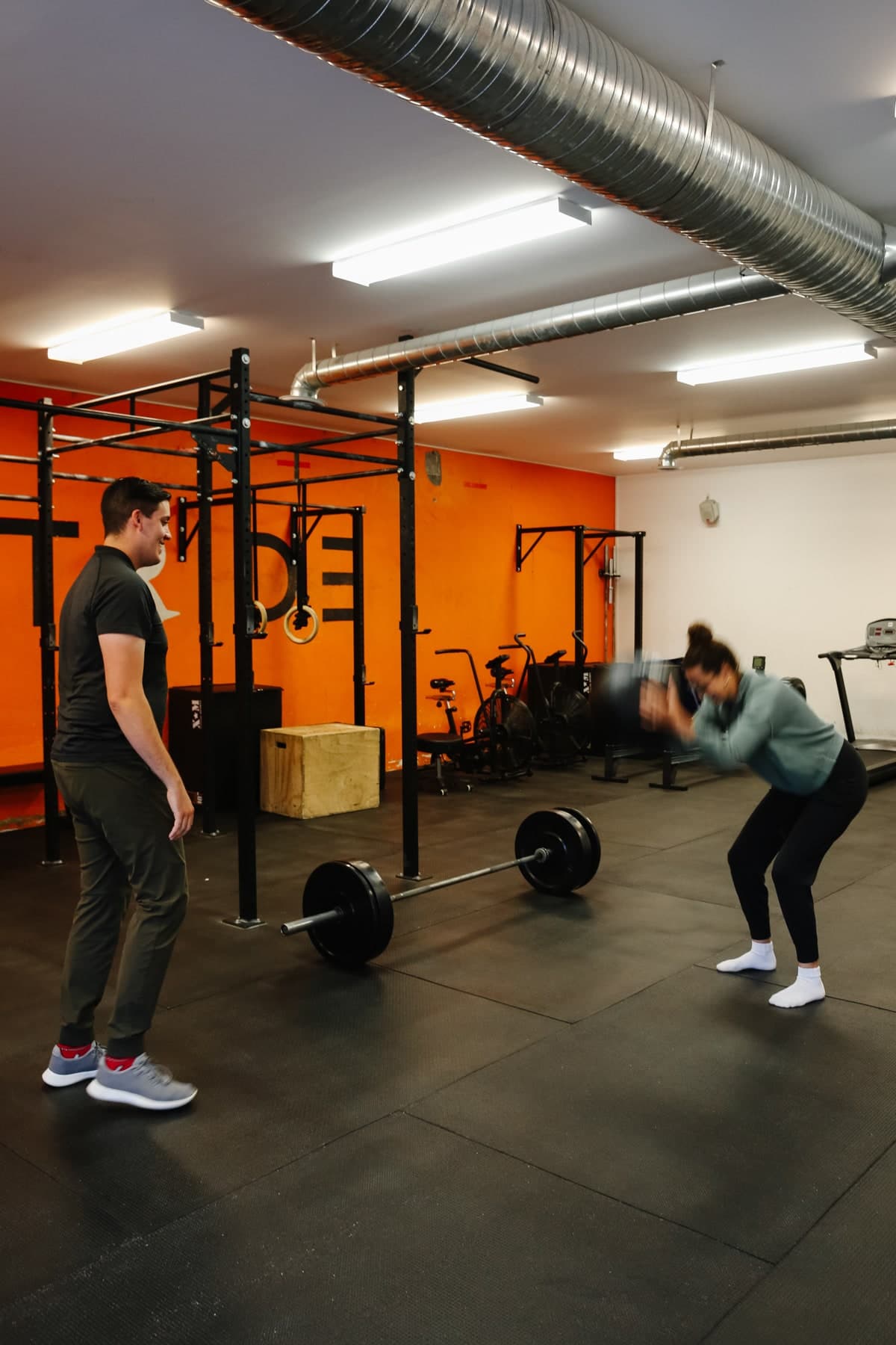 Two people training in a gym with a barbell on the floor, a man in a dark shirt watching and a woman in a teal top squatting with a medicine ball, orange wall, Swift Current
