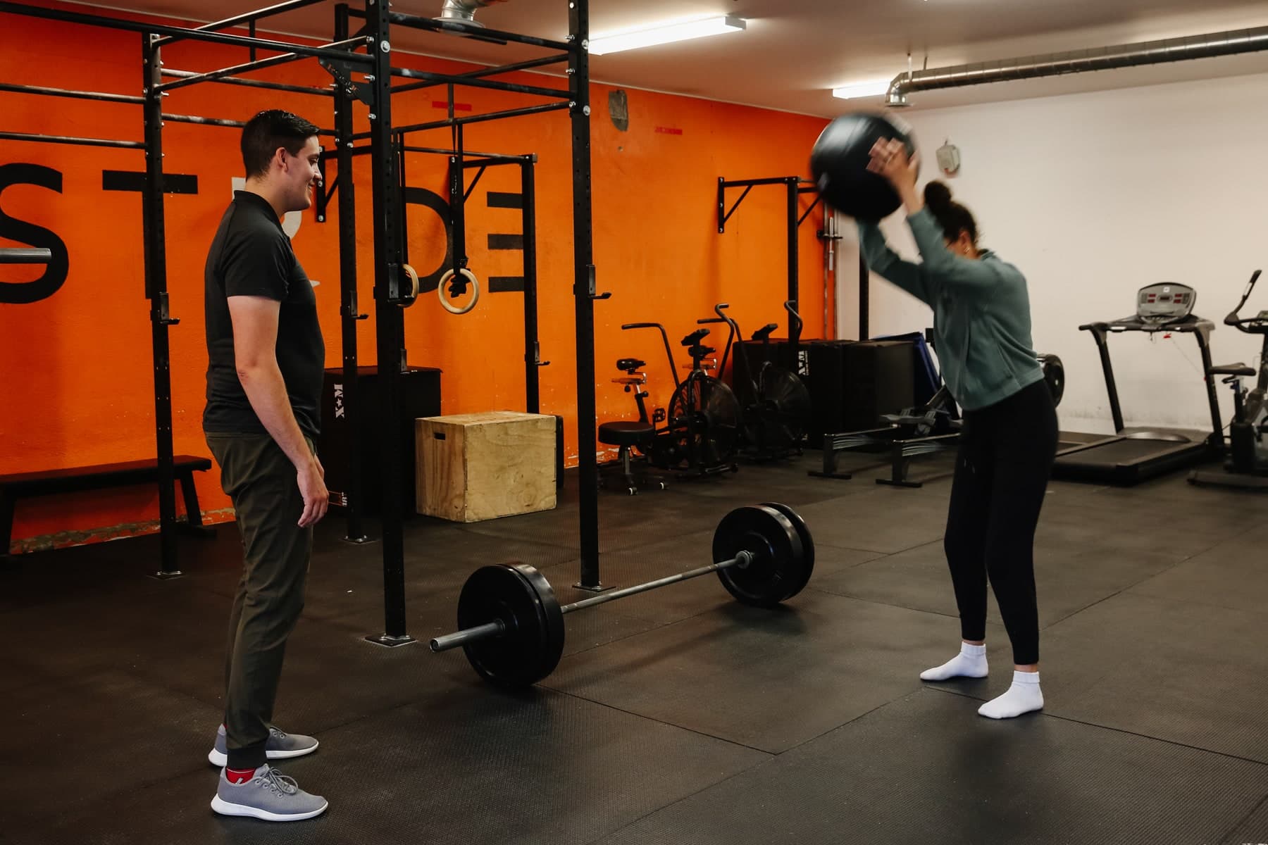 Gym floor with a barbell on black rubber mats and a woman in a grey top throwing a black medicine ball overhead while a man in a dark polo watches, orange wall with black lettering, Swift Current