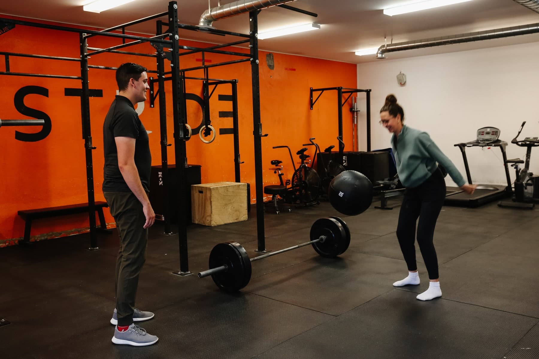 Two people in a gym with a black barbell and medicine ball on the floor, a man in a black shirt watching and a woman in a green sweater smiling and reaching toward the ball, with an orange wall and rig in the background, Swift Current