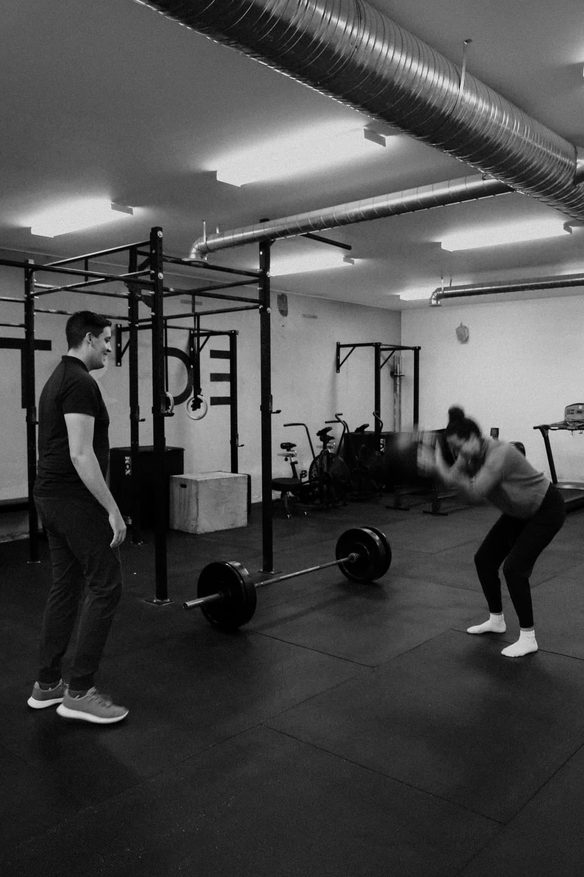 Black and white photo of a gym with a man in a dark polo watching a woman lift a barbell from the floor, rig and ceiling ductwork visible in background, physiotherapist observing exercise in Swift Current