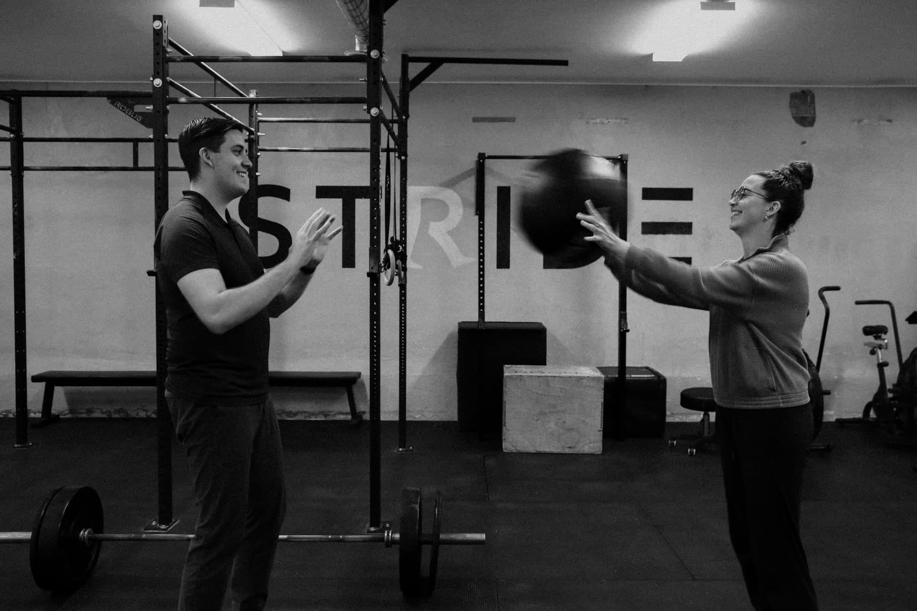 Black and white photo of two people in a gym playing catch with a medicine ball, a man in a dark shirt on the left and a smiling woman with glasses and her hair in a bun on the right, with a barbell and rack behind them and STRIDE lettered on the wall
