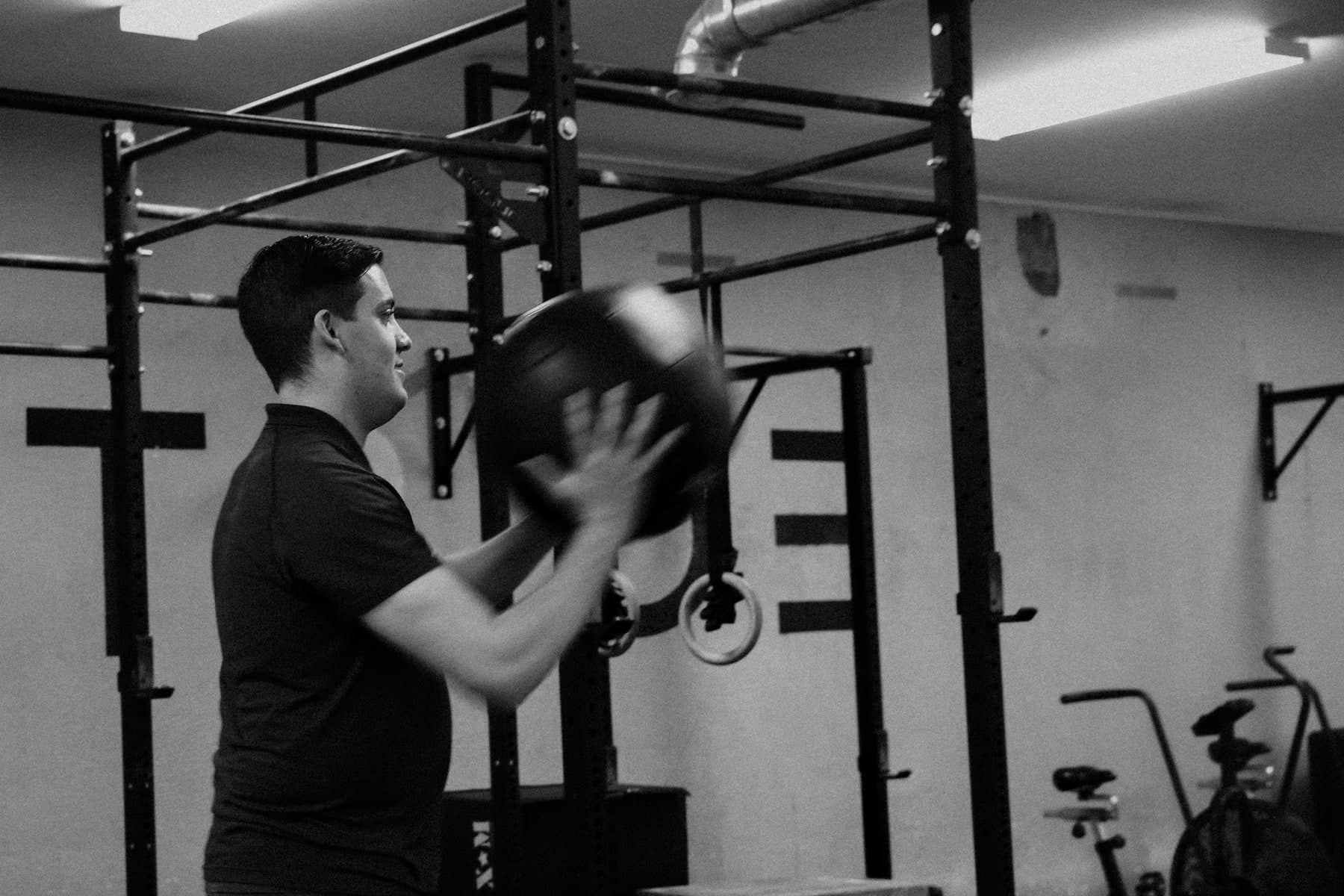 Black and white photo of a man in a dark polo shirt throwing a medicine ball in a gym with a power rack, gymnastic rings and an exercise bike in the background, Swift Current