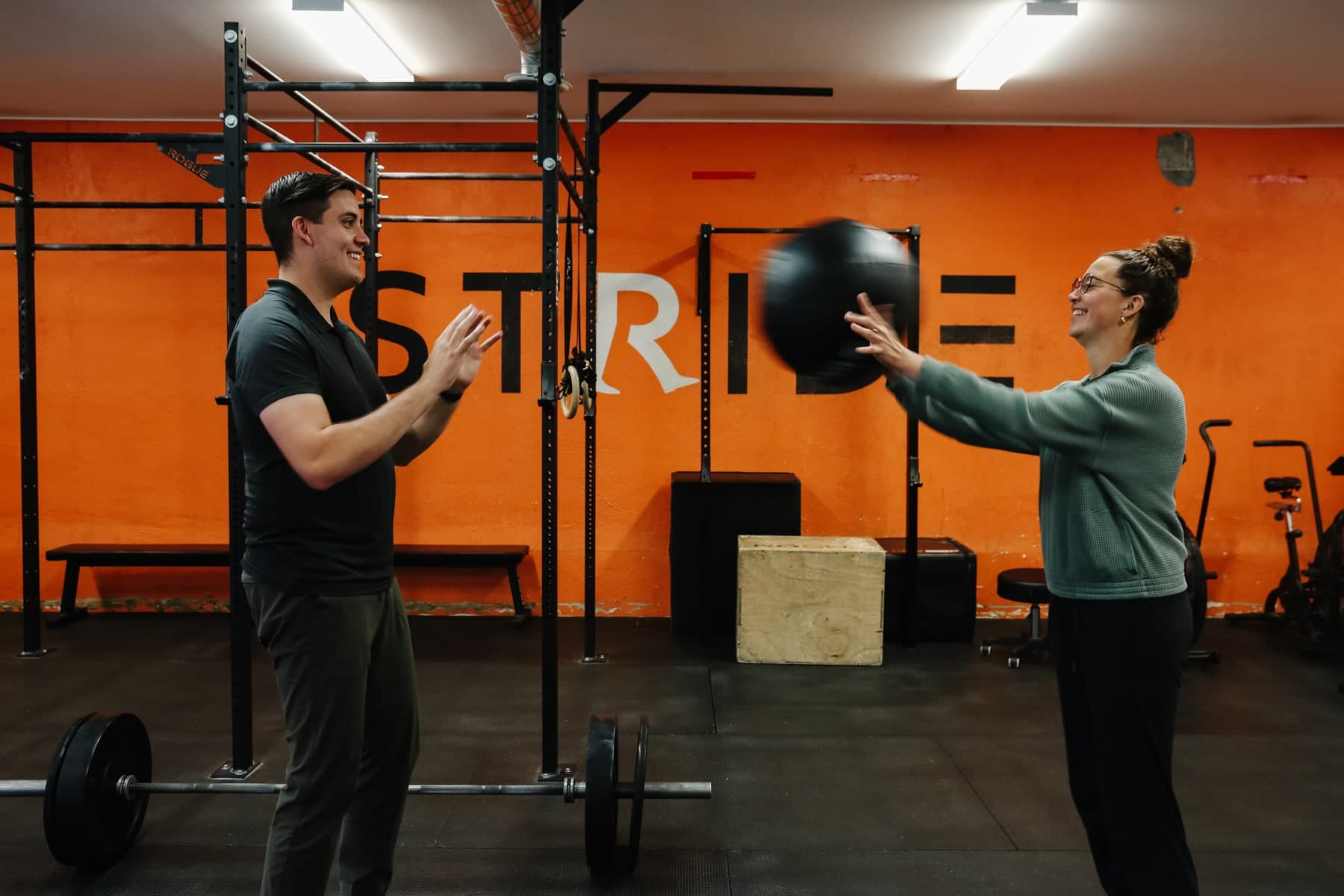 Two people in a gym passing a black medicine ball, a smiling man in a dark polo on the left and a smiling woman in a grey sweater on the right, with a barbell and an orange wall reading STRIDE behind them