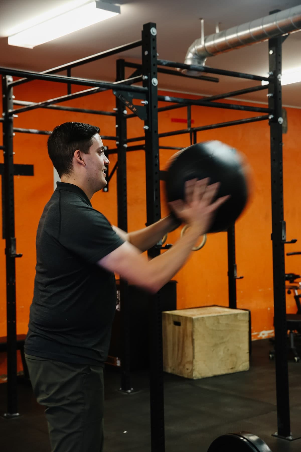 Man in a dark grey polo shirt swinging a black medicine ball in a gym with a black power rack, orange wall and wooden plyo box behind him, Swift Current