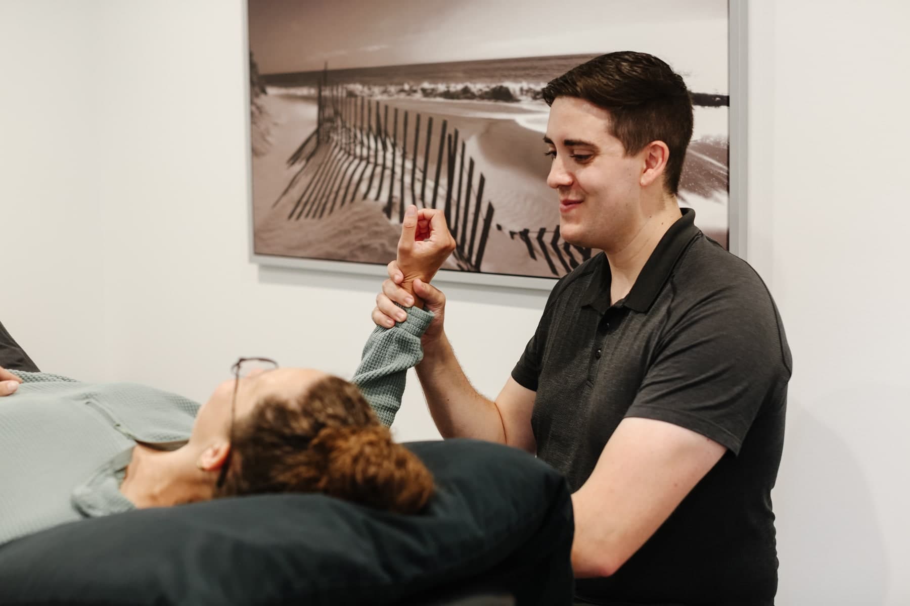Physiotherapist in a dark polo shirt smiling while holding a patient's wrist and hand as the patient lies on a treatment table with a dark pillow, a black and white beach photo with wooden snow fences on the wall behind them