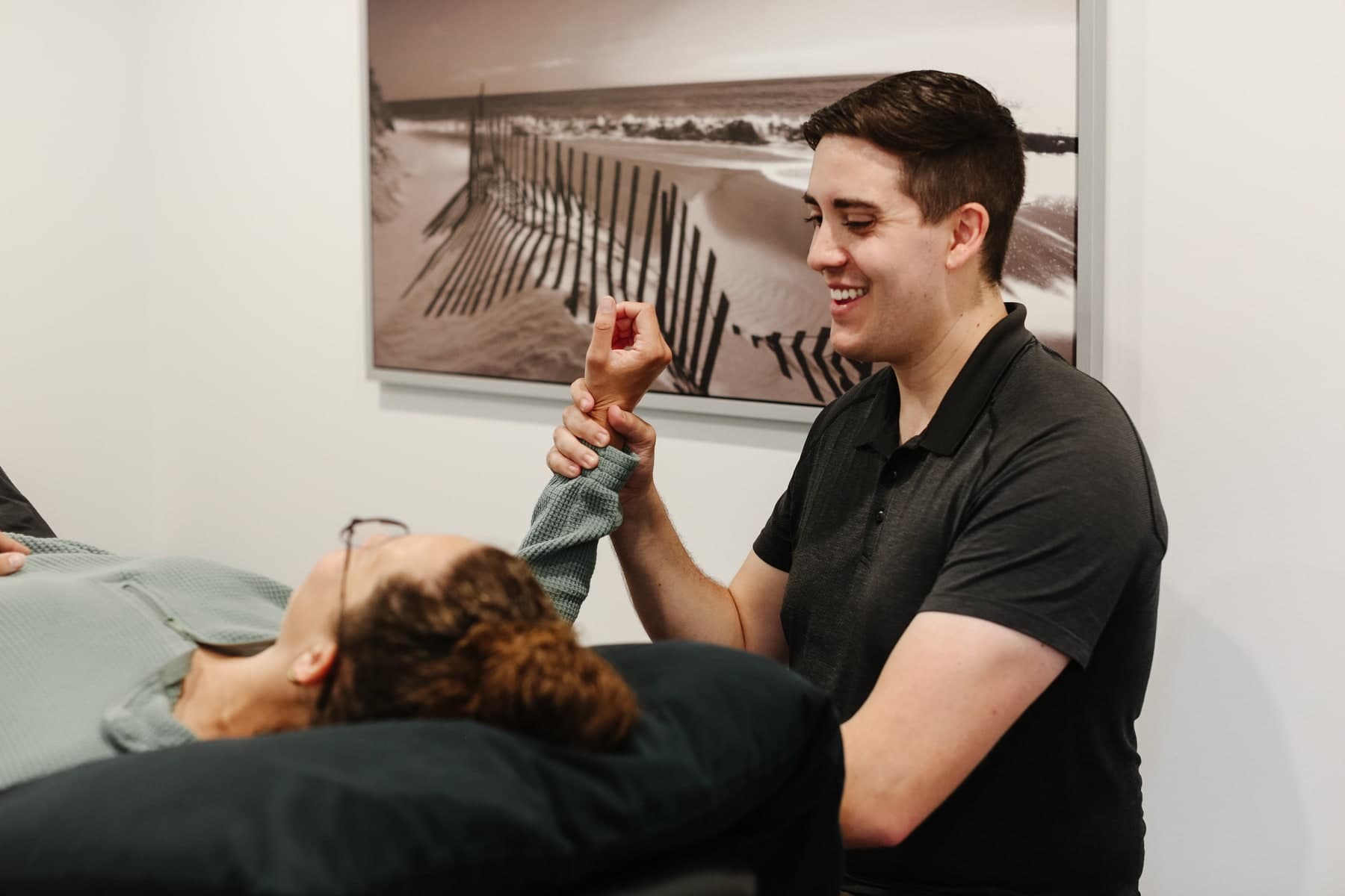 Physiotherapist in a dark polo shirt smiling while gently holding a patient's wrist and forearm as the patient lies on a treatment table, with a black-and-white beach fence photograph on the wall behind