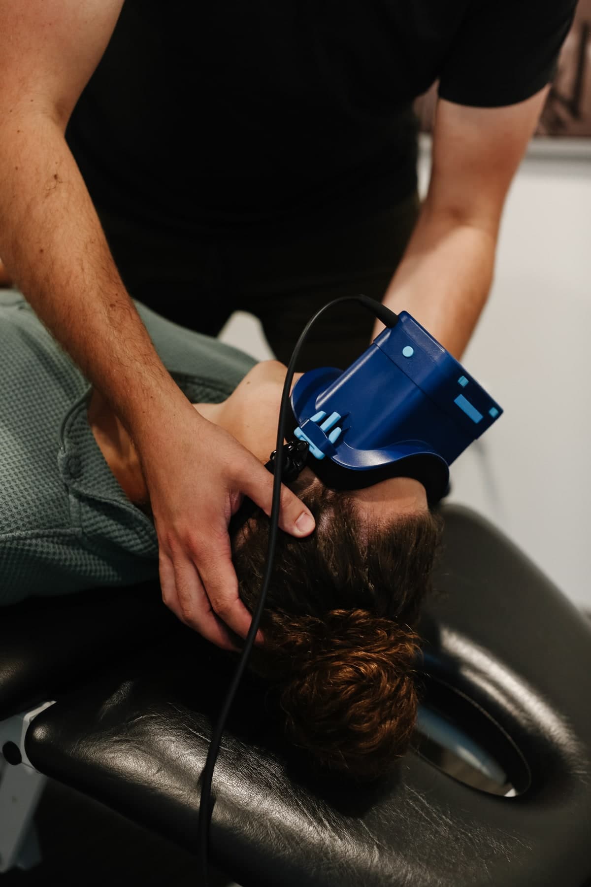 Person lying face-up on a black treatment table with head supported, while a practitioner in a black shirt holds the head and positions a blue handheld device against the neck and base of the skull