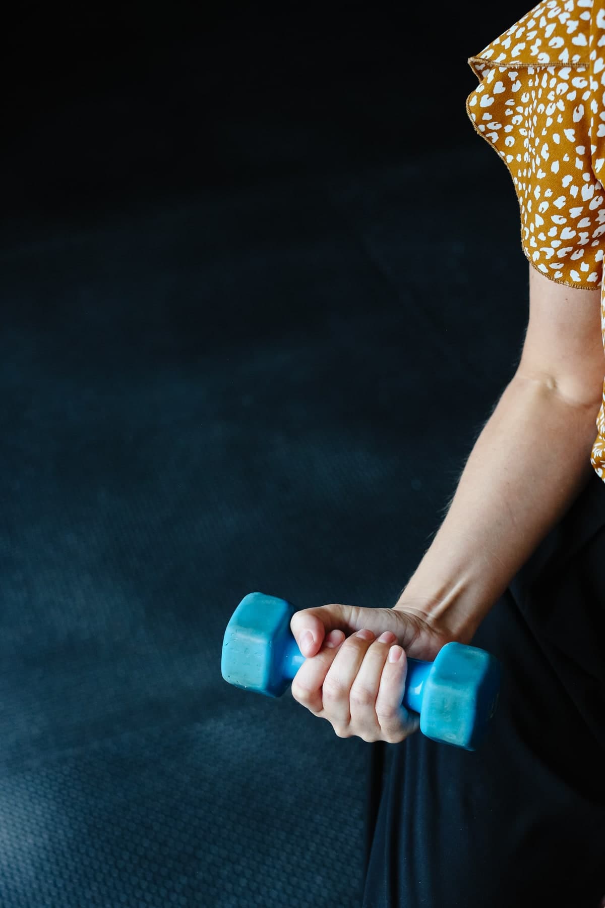 Close-up of a person's hands gripping a blue dumbbell against a dark mat, wearing a mustard top with white spots, in a physiotherapy exercise setting in Swift Current