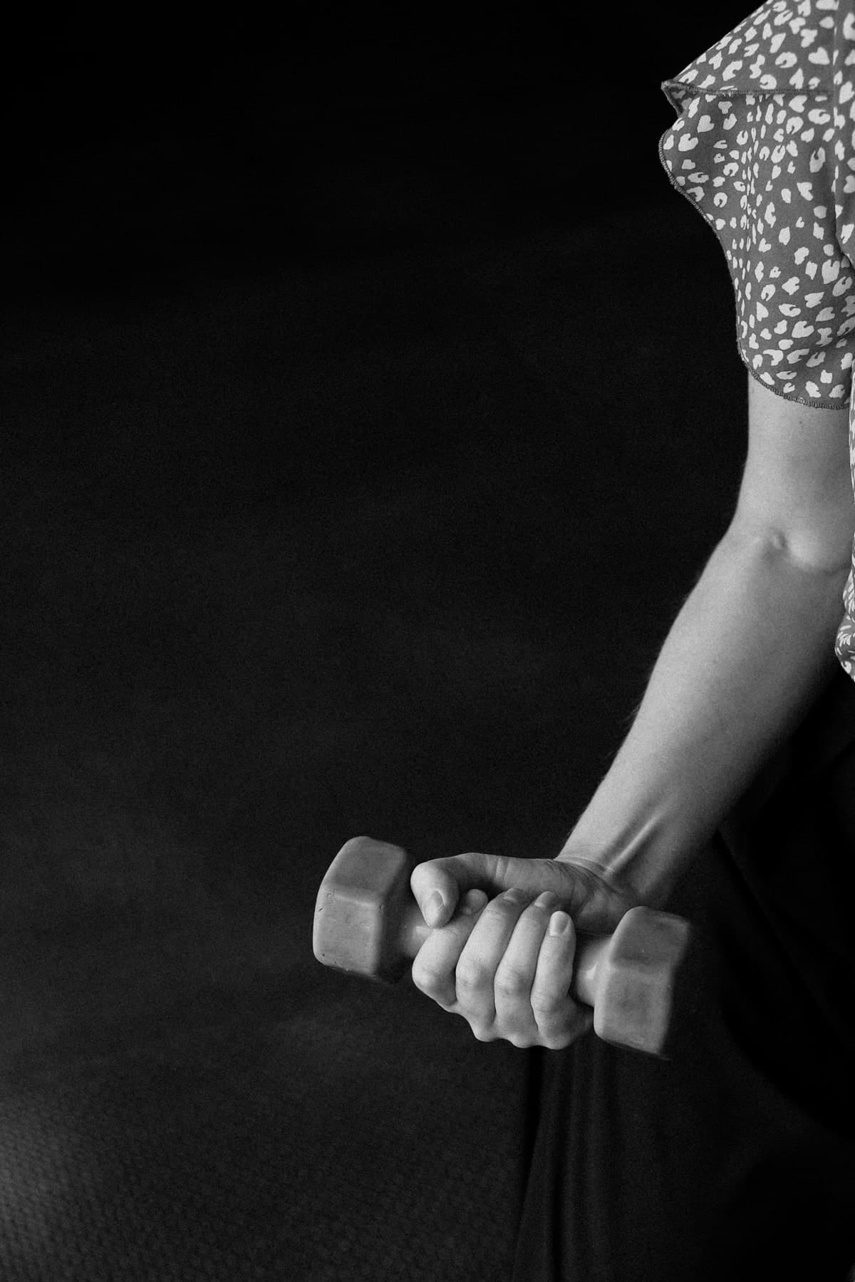 Black and white close-up of a woman's forearm and hand gripping a small hand weight, her arm bent in a biceps curl against a dark background