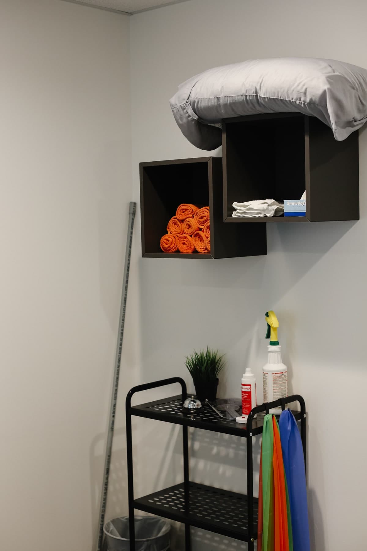Treatment room corner with grey pillow on wall shelves, rolled orange towels, folded white towels, a SnowSoft box, a black utility cart with cleaning spray bottles, a potted plant and colourful resistance bands, Swift Current