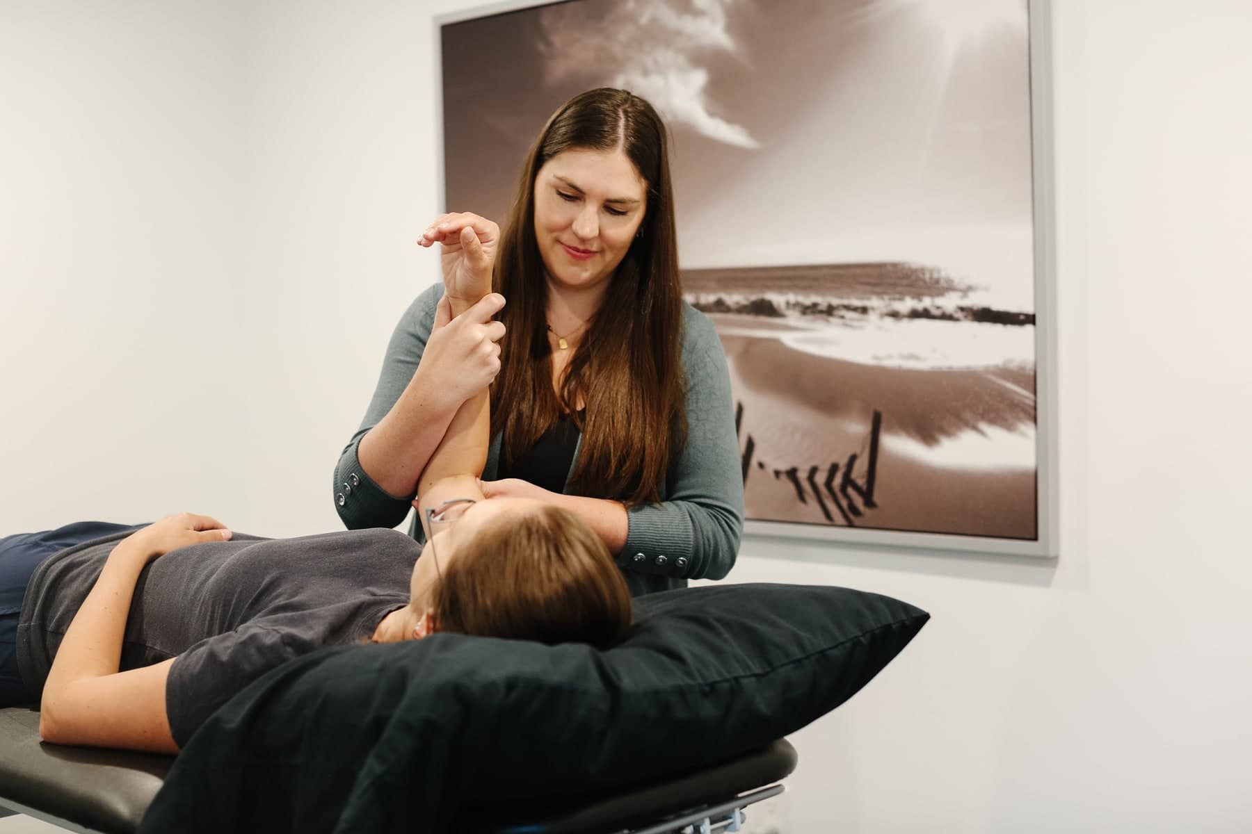 Physiotherapist with long brown hair in a grey-green cardigan gently stretching the arm of a patient lying face up on a treatment table with a dark pillow, framed black-and-white beach photograph on the wall behind, Swift Current