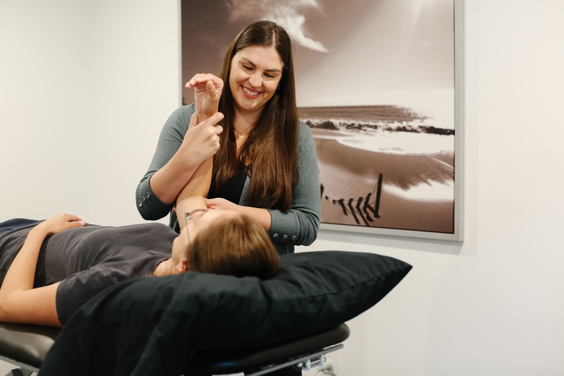 Smiling physiotherapist in a grey cardigan gently stretching the arm of a patient lying on a treatment table in a bright clinic room with a black-and-white beach photograph on the wall