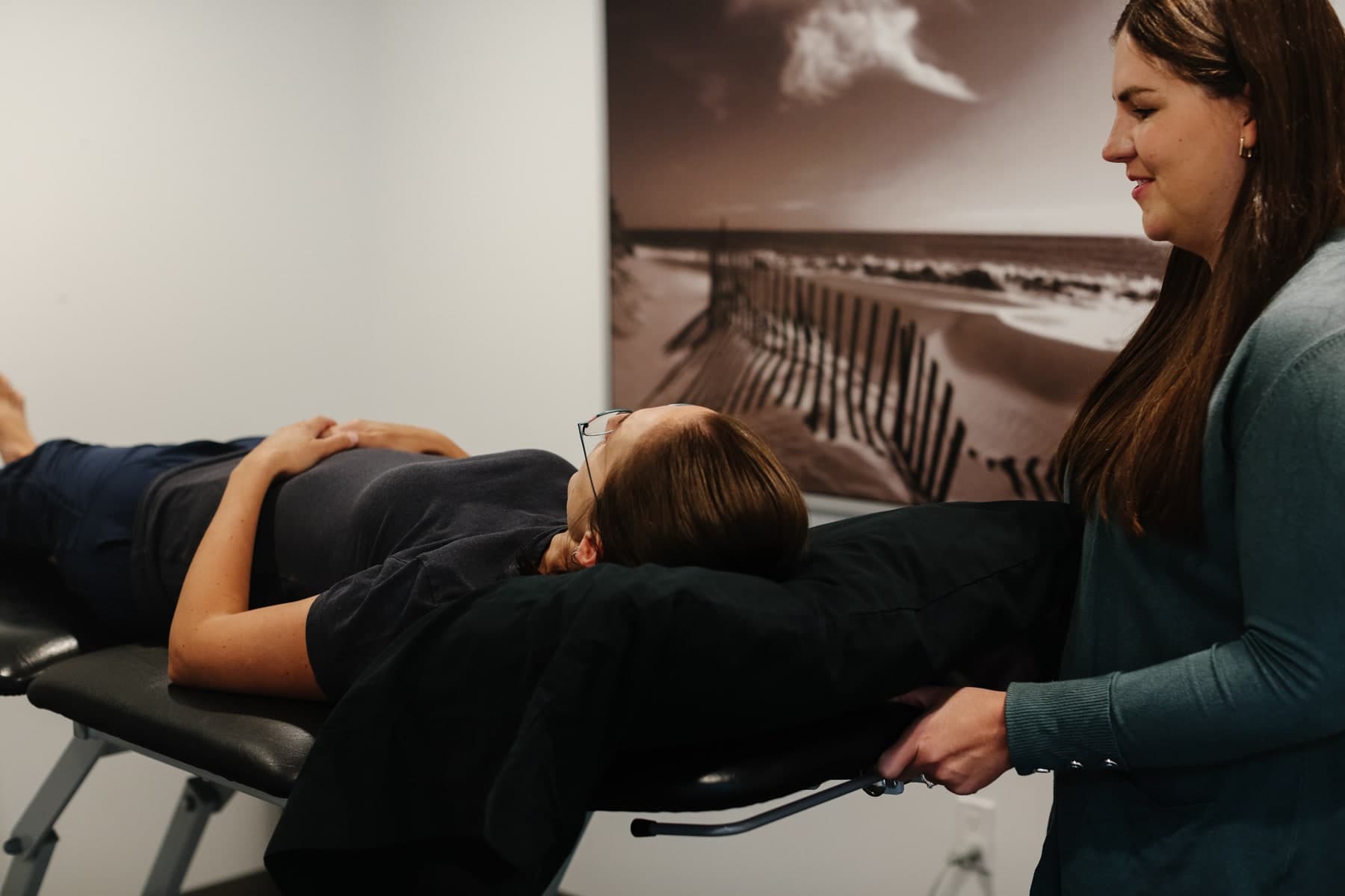 Physiotherapist in a teal cardigan smiling while guiding a patient lying on a black treatment table in a bright clinic room with a sepia beach photograph on the wall, Swift Current