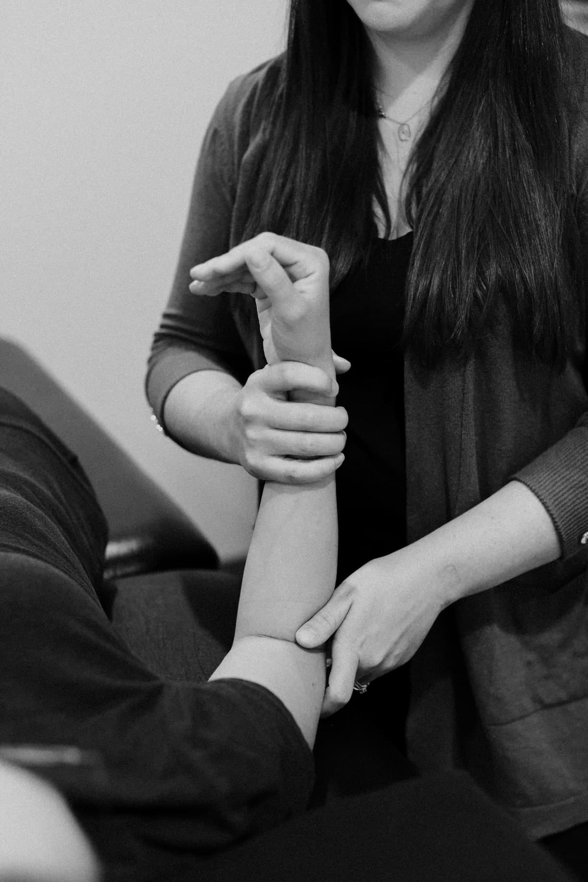 Black and white photo of a physiotherapist with long dark hair in a dark cardigan holding a patient's forearm and wrist, performing a hands-on assessment of the arm, Swift Current