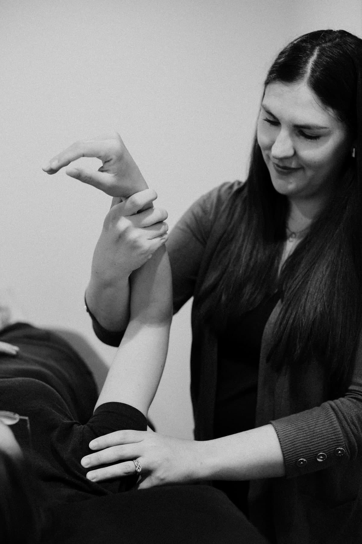 Black and white photo of a smiling female physiotherapist with long dark hair in a dark cardigan gently stretching a patient's raised forearm and wrist while the patient lies back, Swift Current