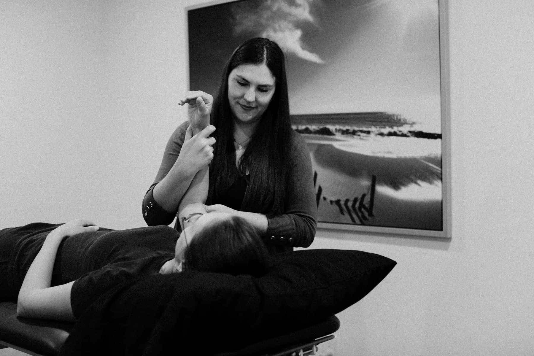 Black and white photo of a physiotherapist with long dark hair holding a patient's raised leg while the patient lies face down on a treatment table with a black pillow, a large framed coastal landscape photograph on the wall behind