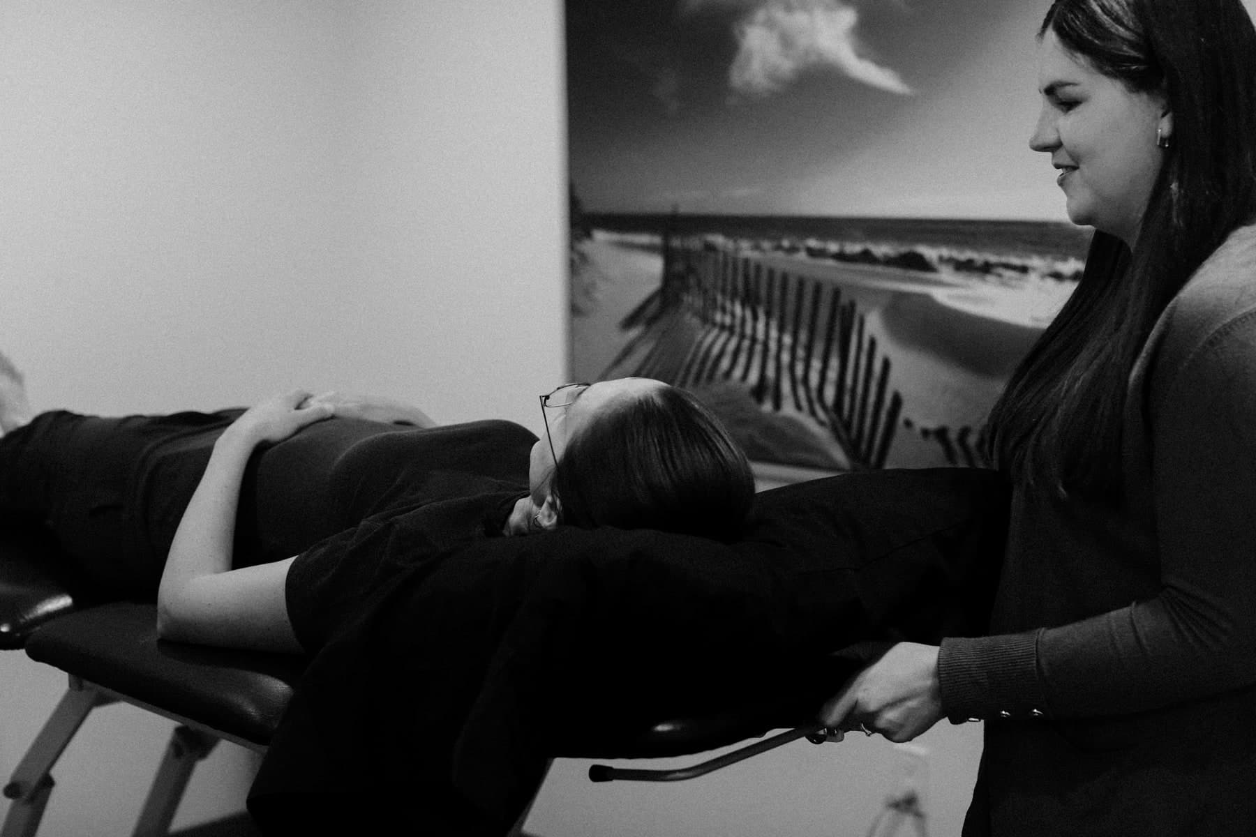 Black and white photo of a patient lying face up on a treatment table while a physiotherapist in a dark sweater stands at the table's end smiling and holding its frame, with a framed beach dune photograph on the wall behind