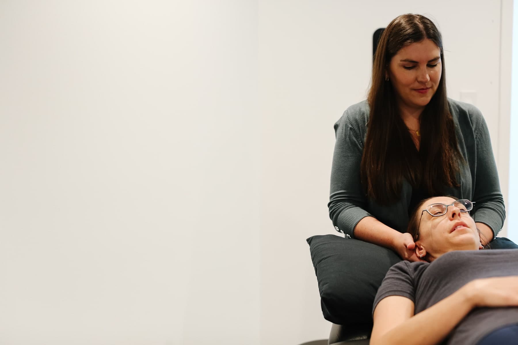 A physiotherapist with long brown hair in a grey cardigan supports the neck of a patient lying on a treatment table with a dark pillow, the patient wearing glasses and a grey top, in a bright white treatment room in Swift Current