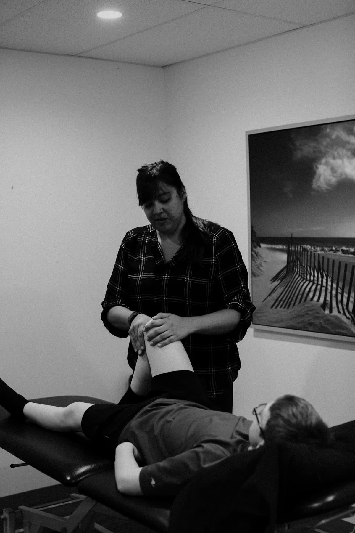 Black and white photo of a physiotherapist in a plaid shirt holding a patient's leg while the patient lies on a treatment table in a clinic room with a framed beach photograph on the wall, Swift Current