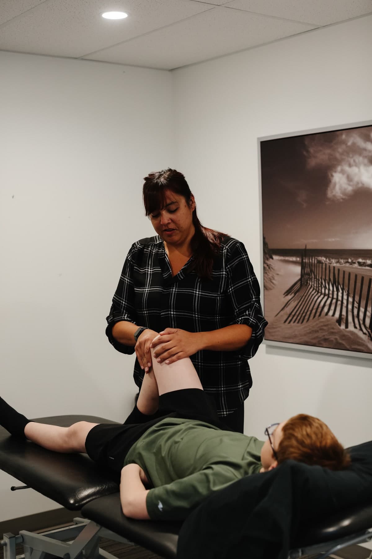 Physiotherapist in a black and white plaid blouse holding a patient's lower leg while the patient lies on a treatment table, framed sepia photograph of a beach with wooden fencing on the wall, Swift Current