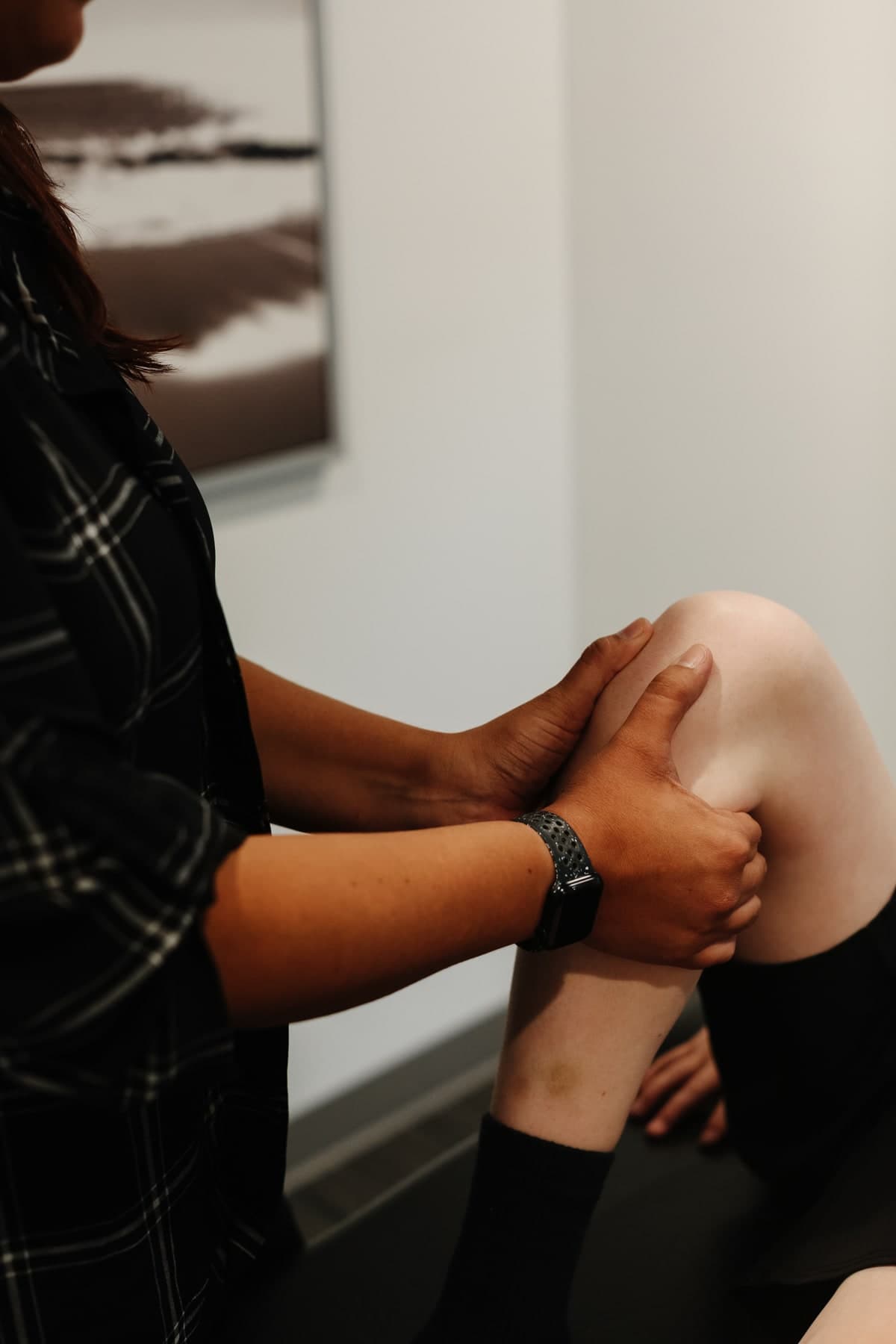A physiotherapist in a black plaid top with her hands on a patient's knee, examining or treating the joint in a clinic room with a framed black and white landscape photo on the wall