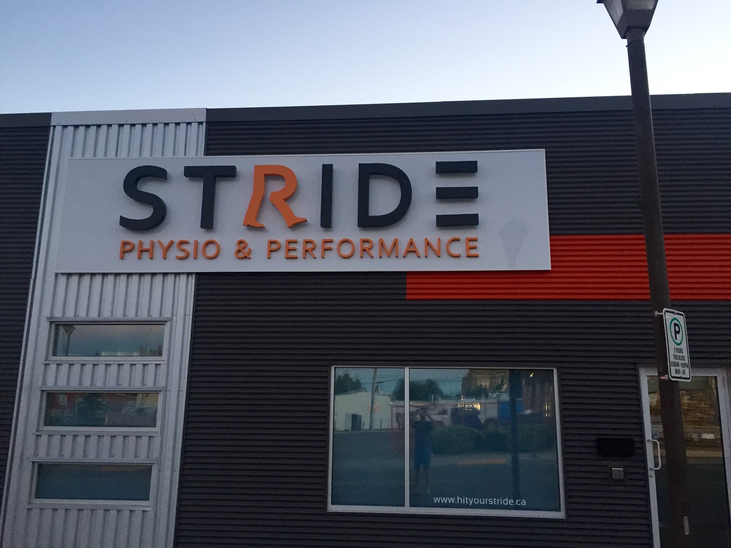 Exterior sign reading STRIDE with PHYSIO & PERFORMANCE beneath, dark letters with an orange R, on a dark corrugated building with an orange band and a window showing the website address