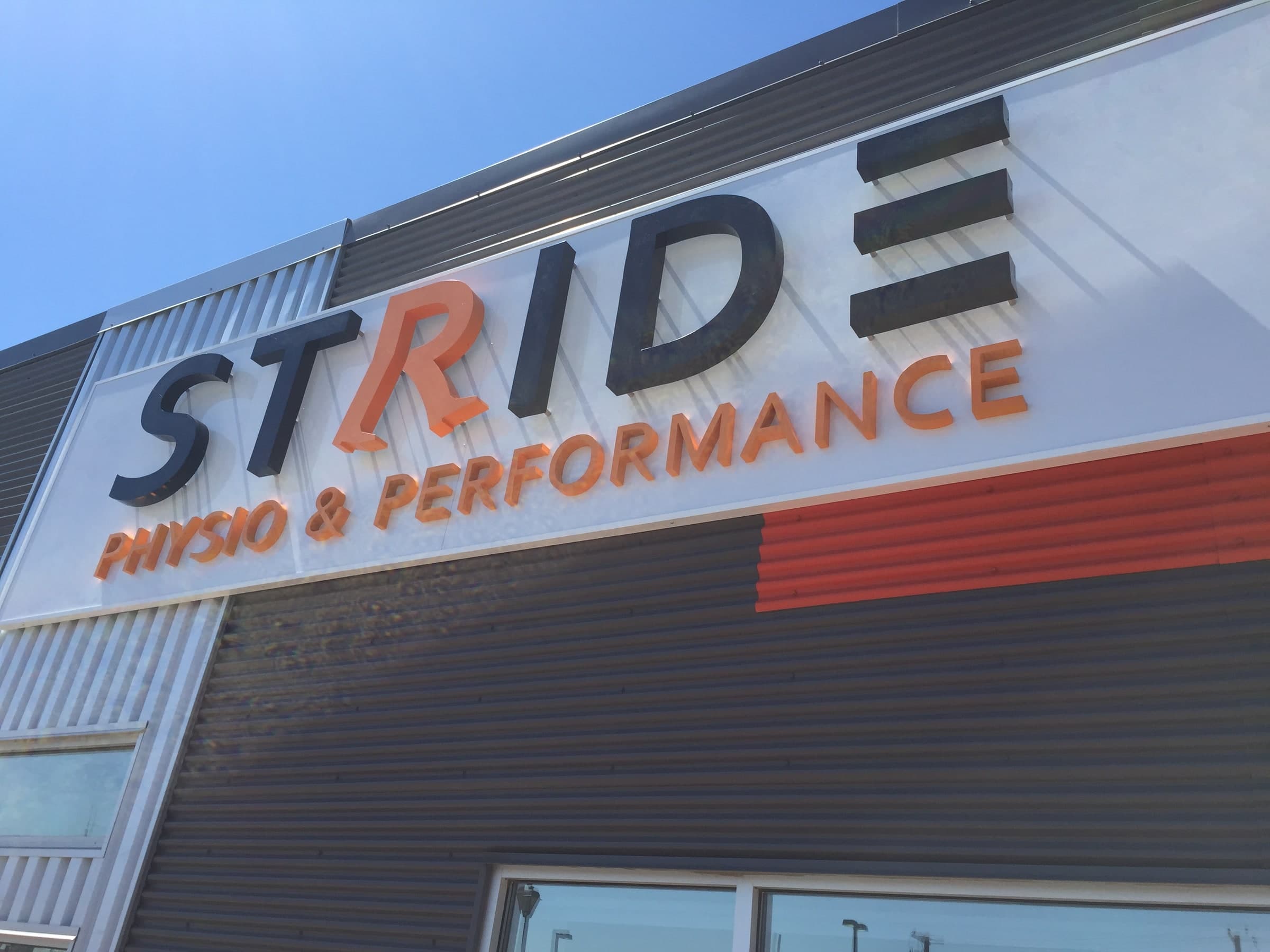 Exterior building sign reading STRIDE with PHYSIO & PERFORMANCE beneath it in orange and dark grey lettering on a white panel above corrugated metal cladding