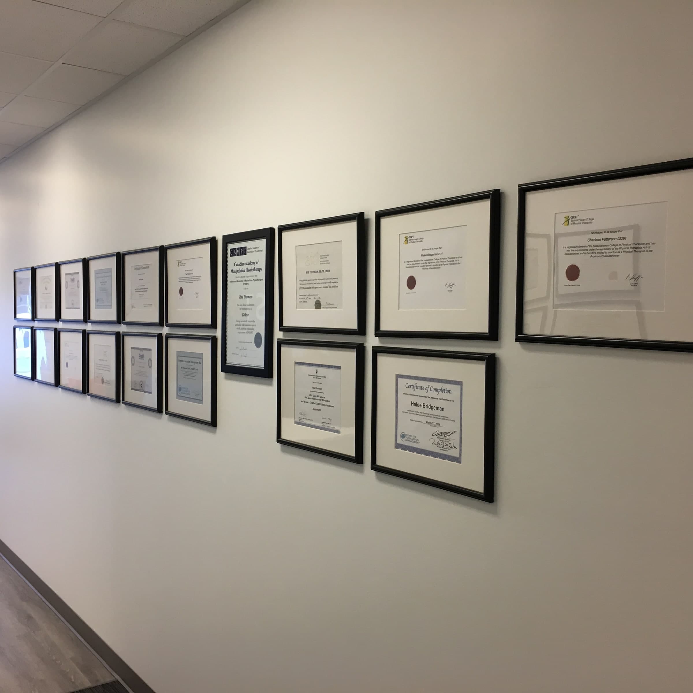 Hallway wall lined with framed professional certificates and credentials, including a Canadian Academy of Manipulative Physiotherapy fellowship certificate and Saskatchewan College of Physical Therapists registration certificates, in Swift Current