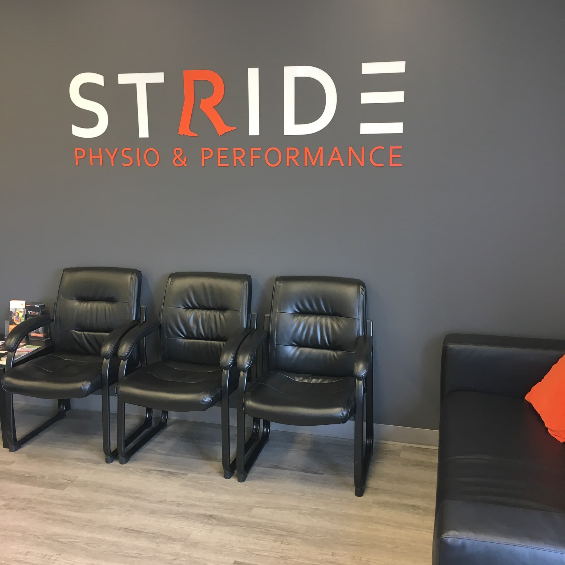 Waiting area with three black leather chairs against a grey wall displaying the white and orange STRIDE PHYSIO & PERFORMANCE sign, and a black sofa with an orange cushion at right
