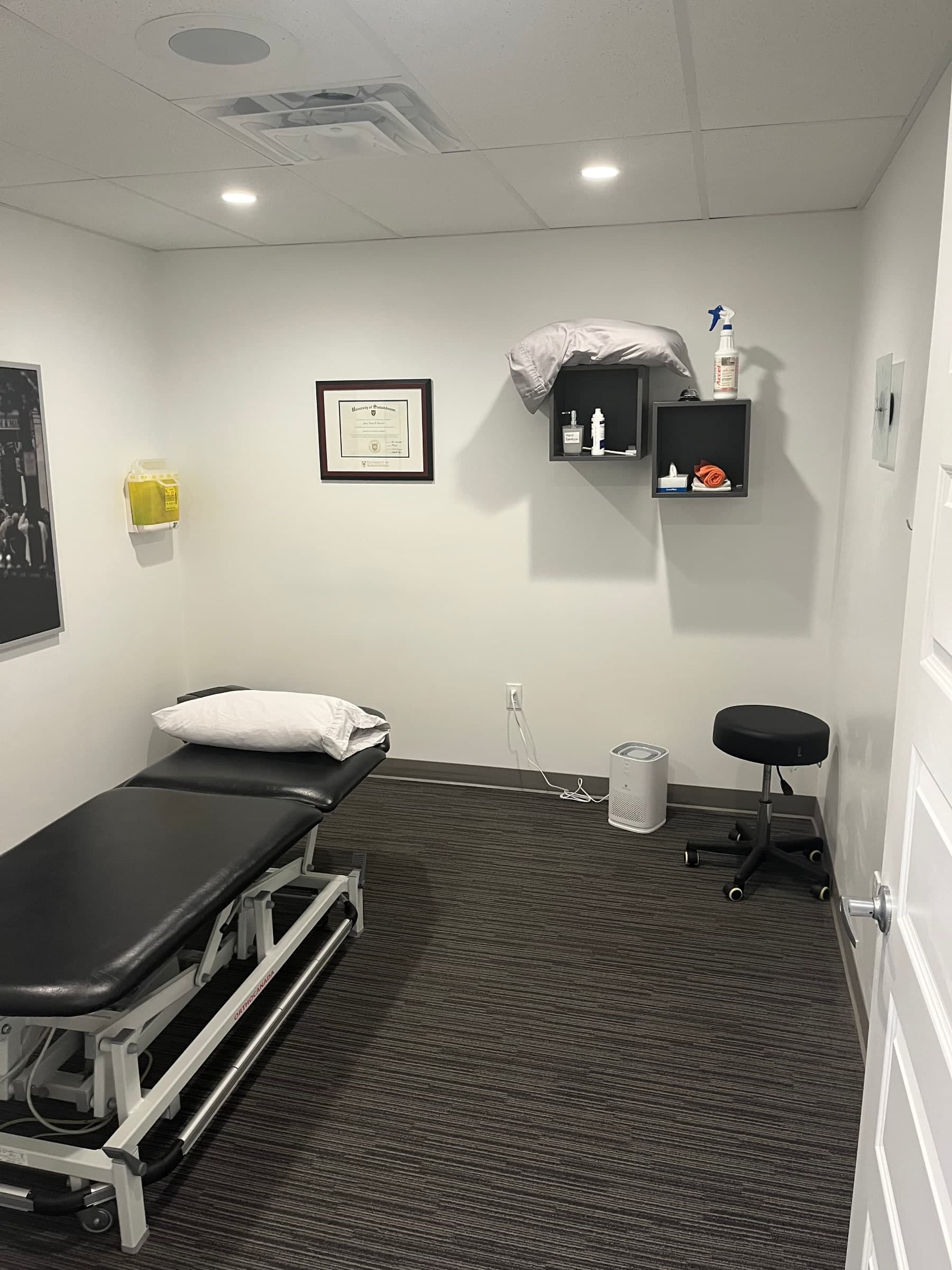 Treatment room with a black and white adjustable treatment table with a pillow, a black rolling stool, a framed University of Saskatchewan certificate on the wall, wall shelves with hand sanitizer and supplies, and an air purifier on the floor, Swift Current