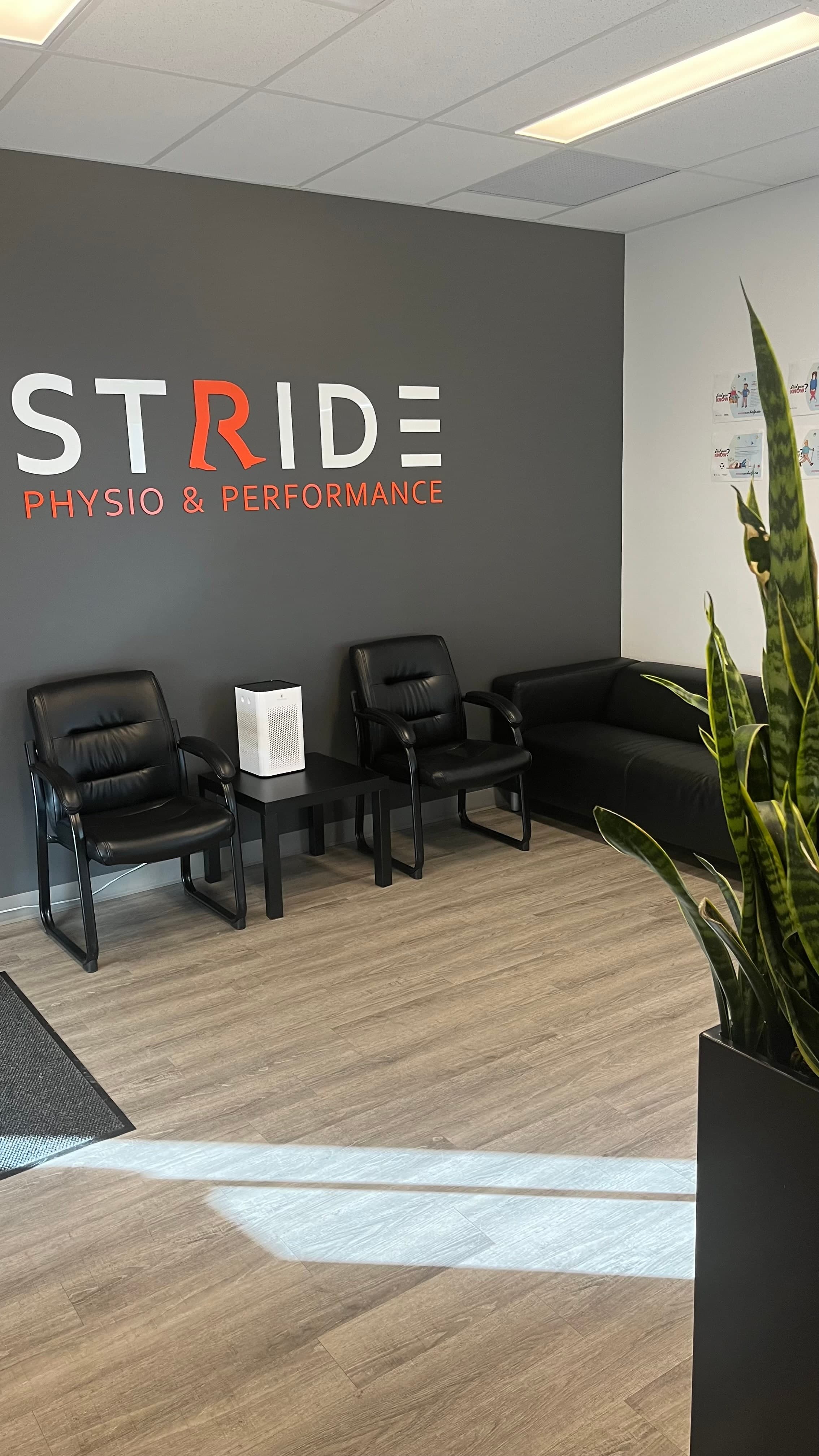 Waiting area with black leather chairs and a black sofa in front of a dark grey wall with the Stride Physio & Performance logo in white and orange letters, a white air purifier on a side table, and a snake plant in a black planter