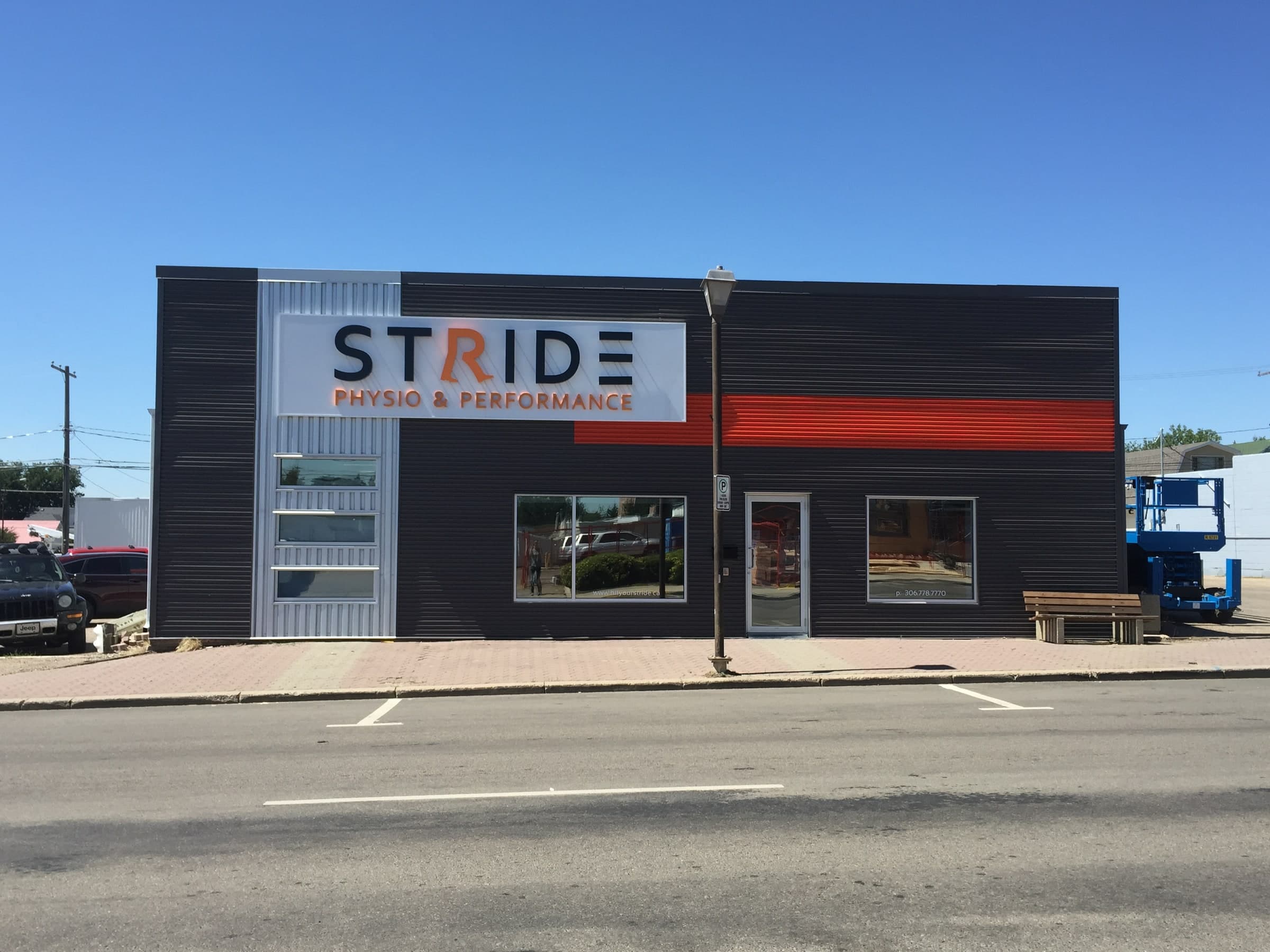 Exterior of Stride Physio and Performance clinic showing the illuminated STRIDE sign with PHYSIO & PERFORMANCE beneath it, a dark corrugated facade with an orange stripe, and a blue scissor lift beside the building in Swift Current