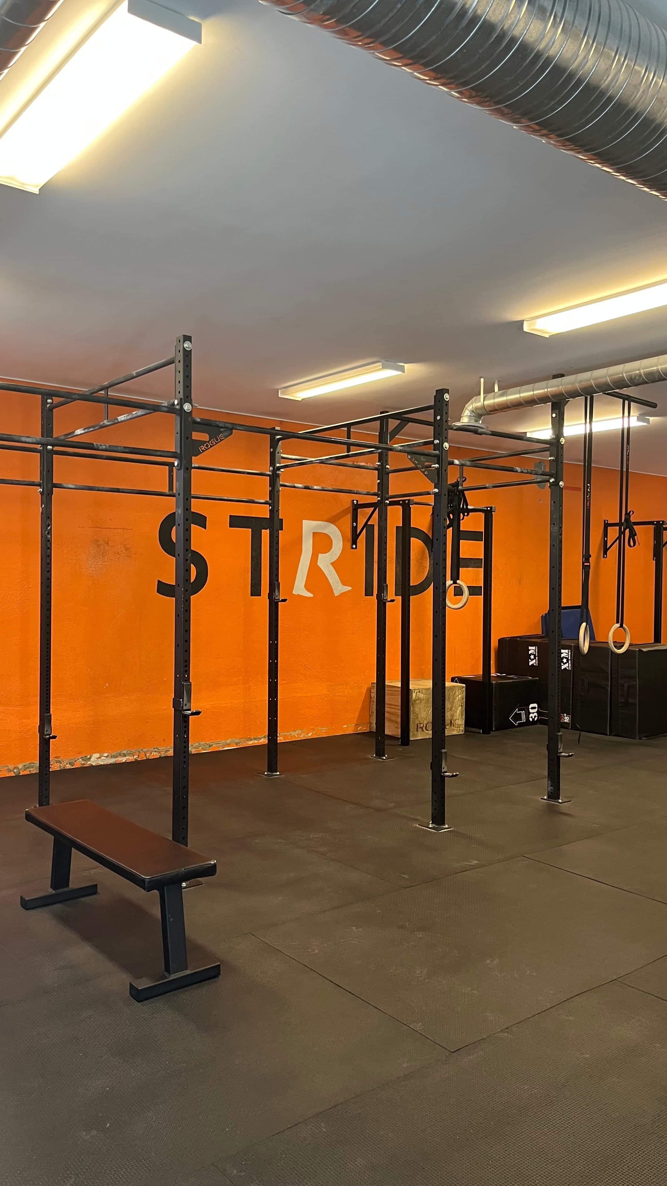 Interior of a functional training gym with black power racks and pull-up bars, gymnastic rings hanging on the right, wooden plyometric boxes, a flat bench in the foreground, and a bright orange wall with large STRIDE lettering in Swift Current